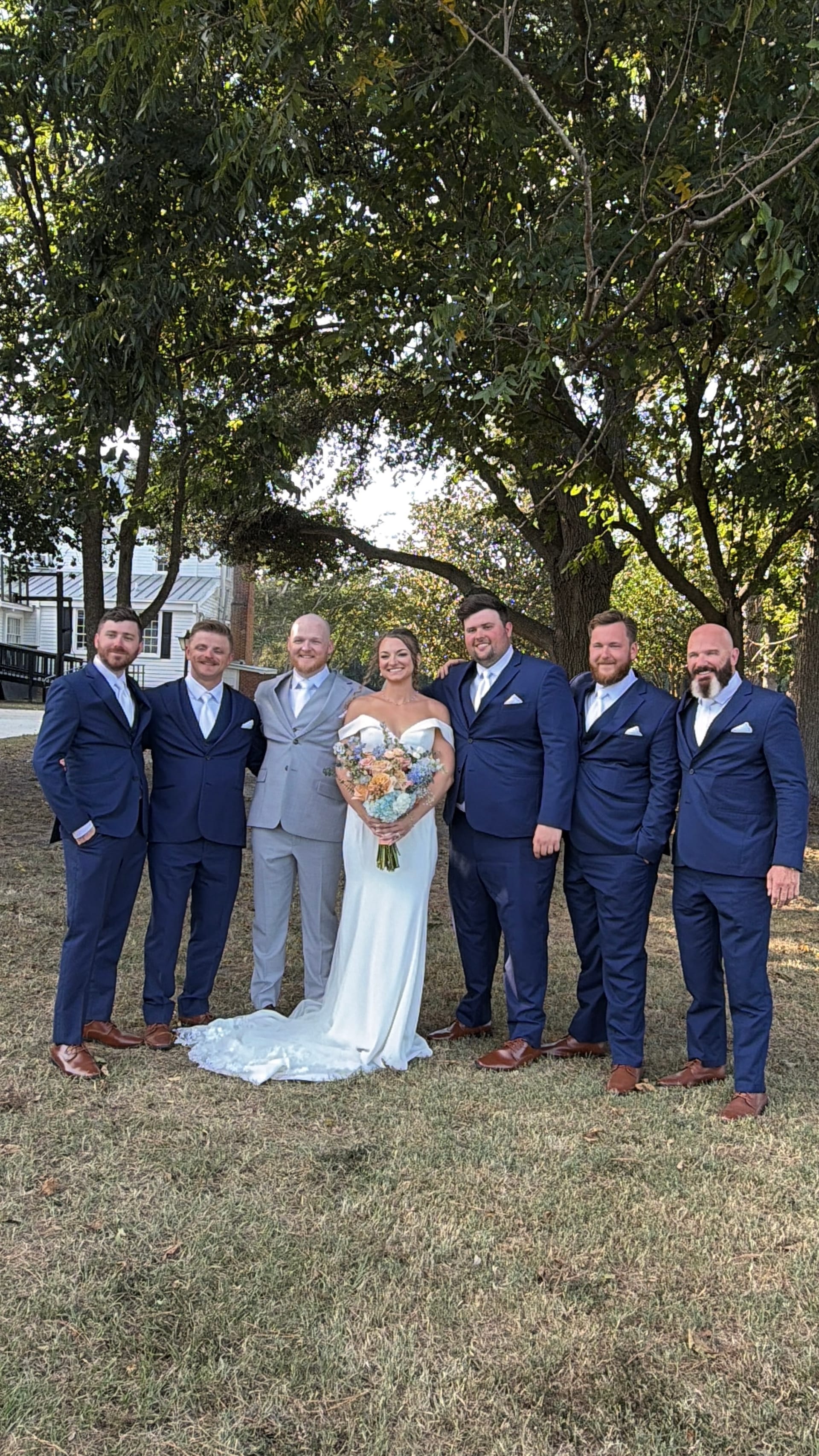 Bride in an off-shoulder white gown holding a pastel bouquet stands with five groomsmen in navy suits beneath the sweeping oak trees at Scarborough House.