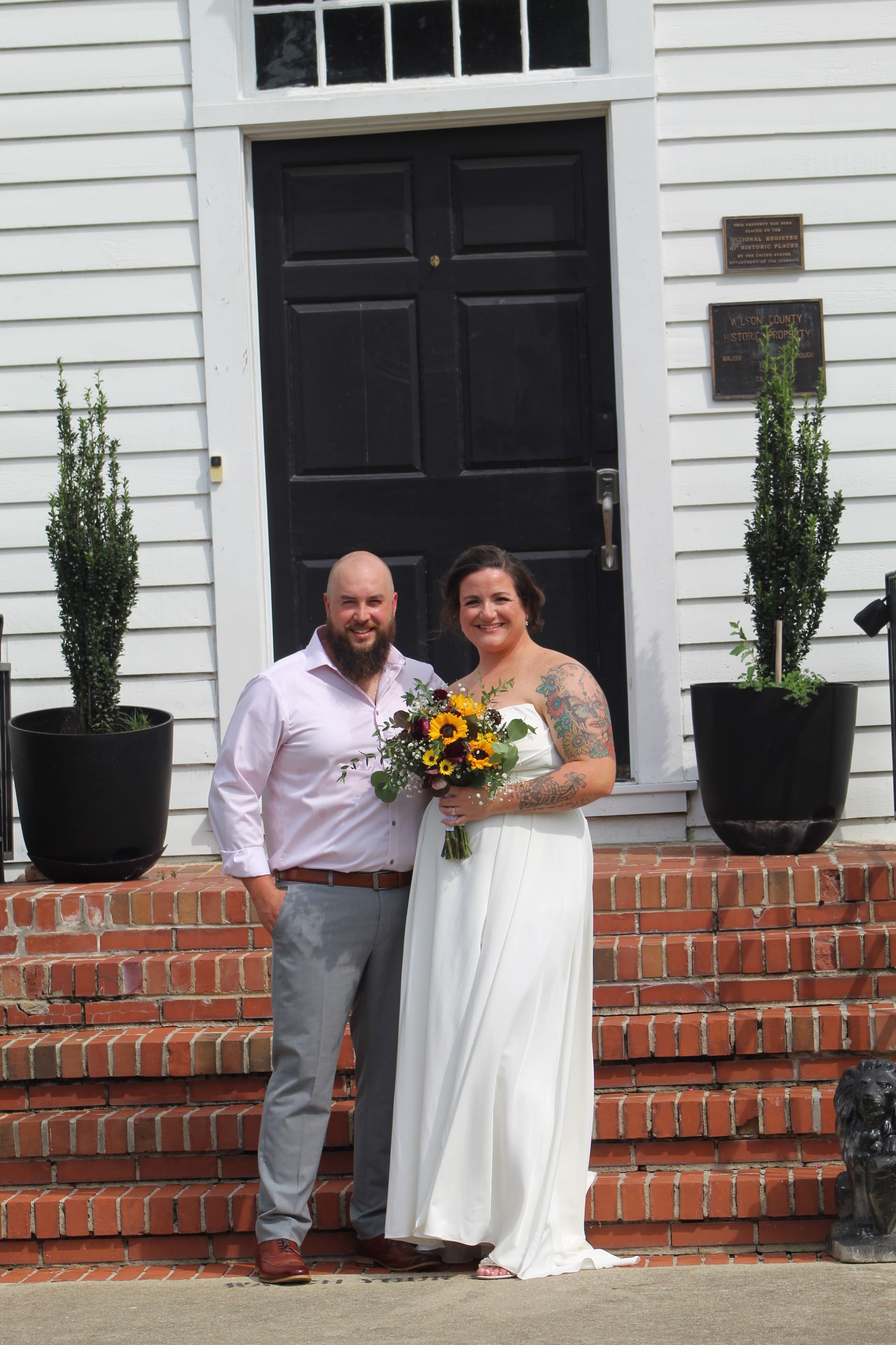 Bride in a white gown holding a sunflower bouquet stands with her bearded groom in a pink dress shirt on the brick steps of the historic 1821 Scarborough House entrance.