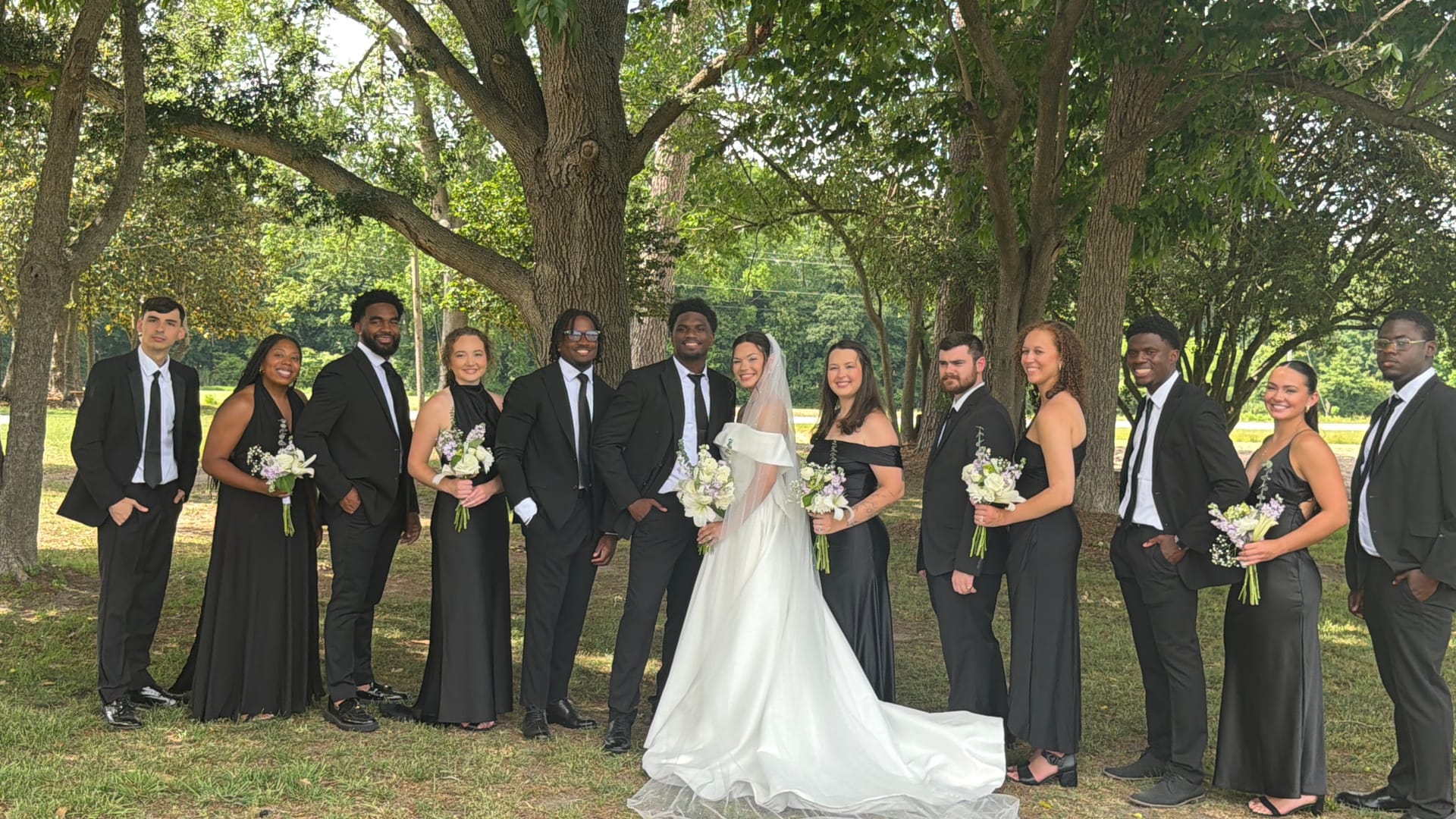 Bride in white gown and groom in black suit pose with their wedding party dressed in all black beneath sprawling oak trees at Scarborough House estate.