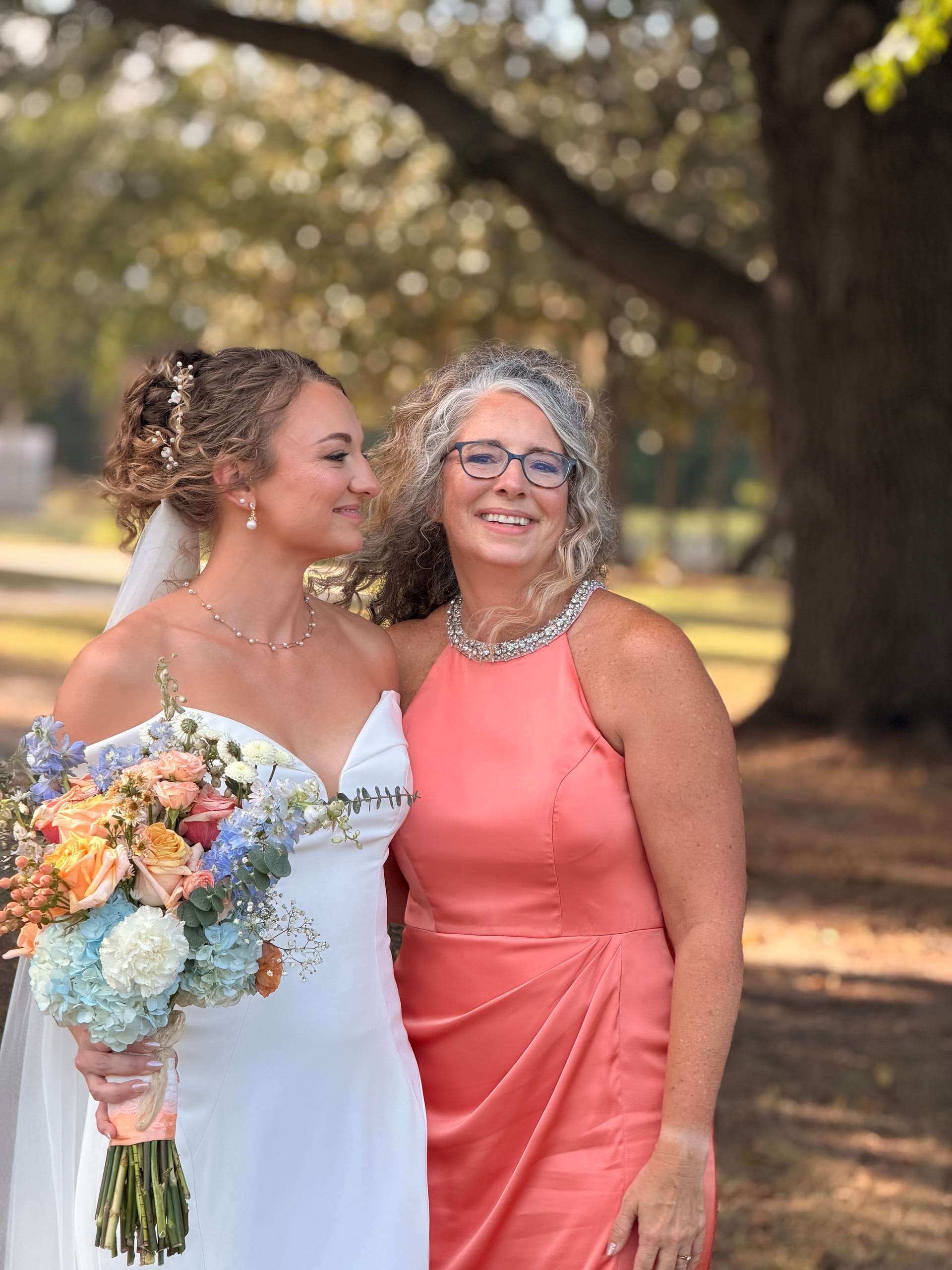 Bride in a white gown holding a pastel bouquet smiles with an older woman in a coral dress beneath the sweeping oak trees at Scarborough House.