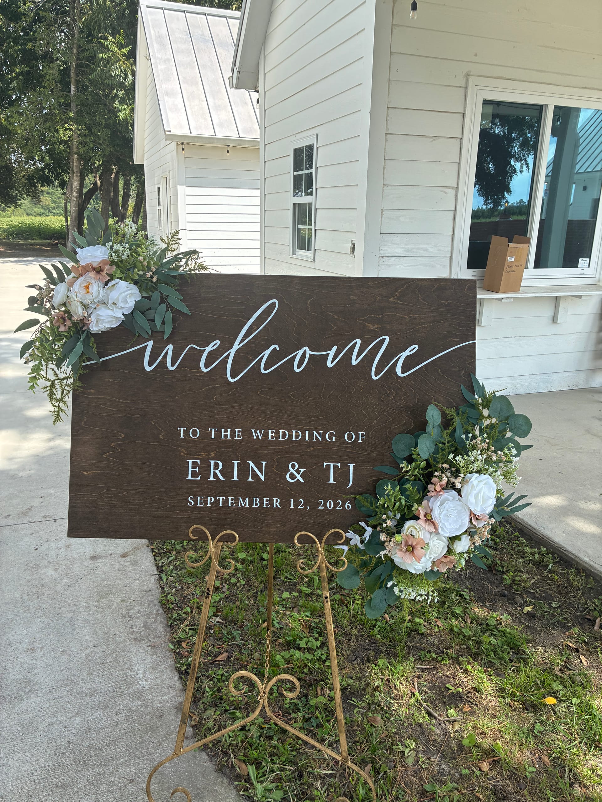 Rustic dark-wood wedding welcome sign for Erin and TJ on September 12 2026 adorned with white and blush floral arrangements on a gold easel outside a white historic farmhouse venue.