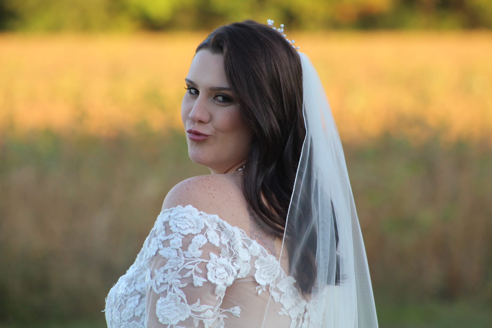 Bride in an off-shoulder lace gown and white veil glances over her shoulder with a soft smile during a golden-hour portrait in a sunlit field at Scarborough House.
