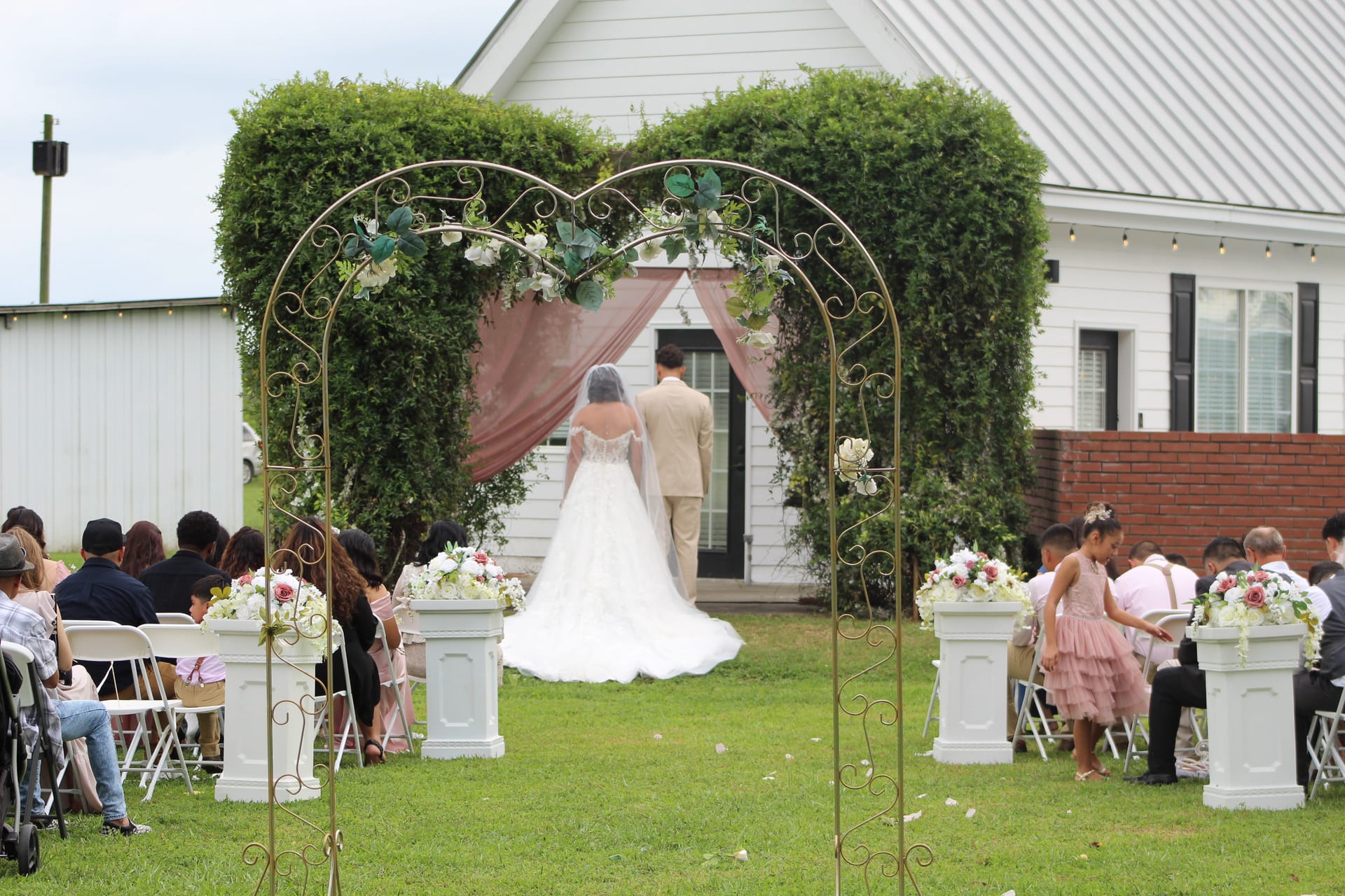 Bride and groom stand at a gold scrollwork arch adorned with white flowers and blush drapery on the ceremony lawn at Scarborough House, surrounded by seated guests on a lush green estate.