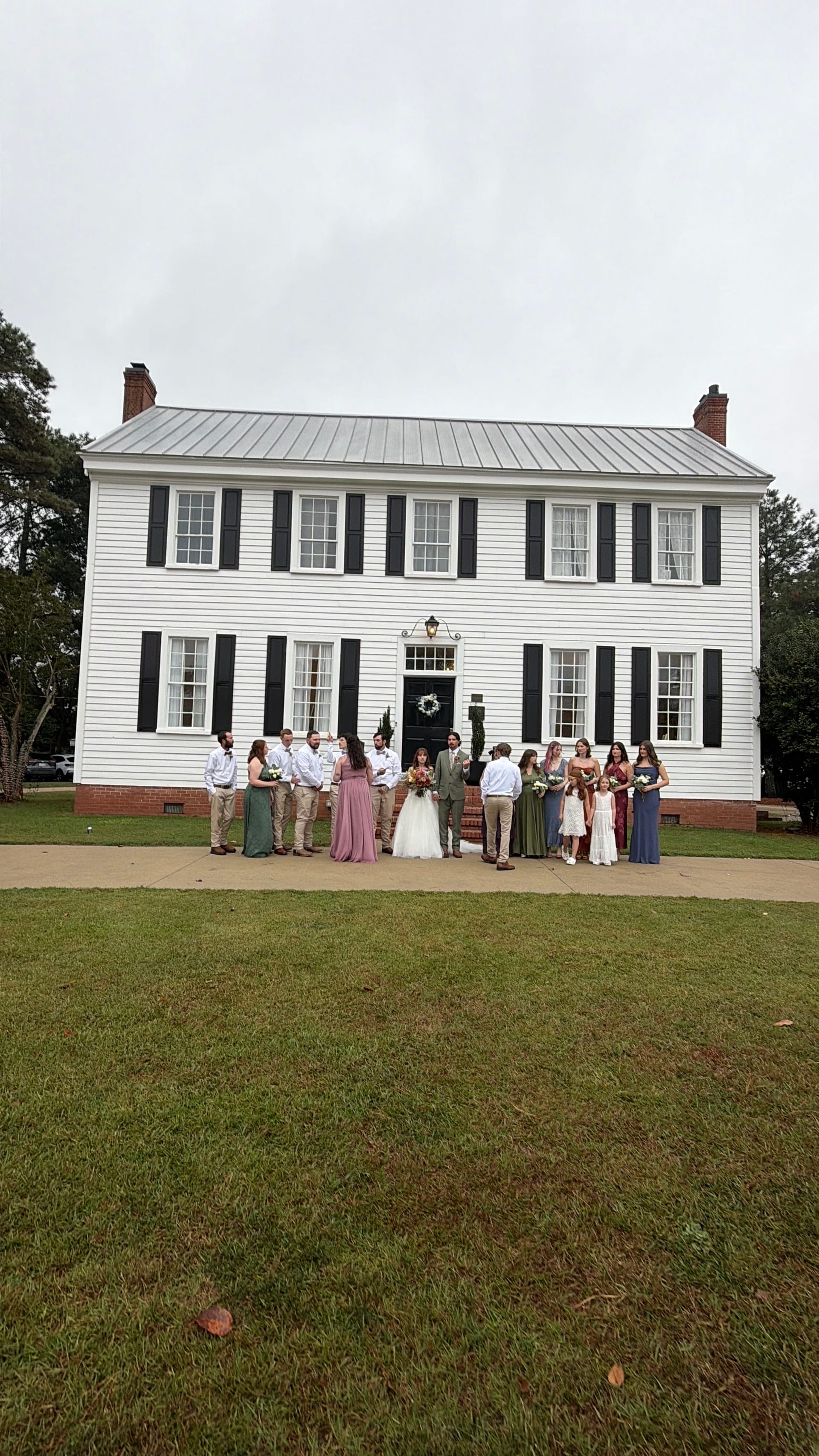 Full wedding party posed in front of the white clapboard 1821 Scarborough House manor with black shutters and metal roof on an overcast day.