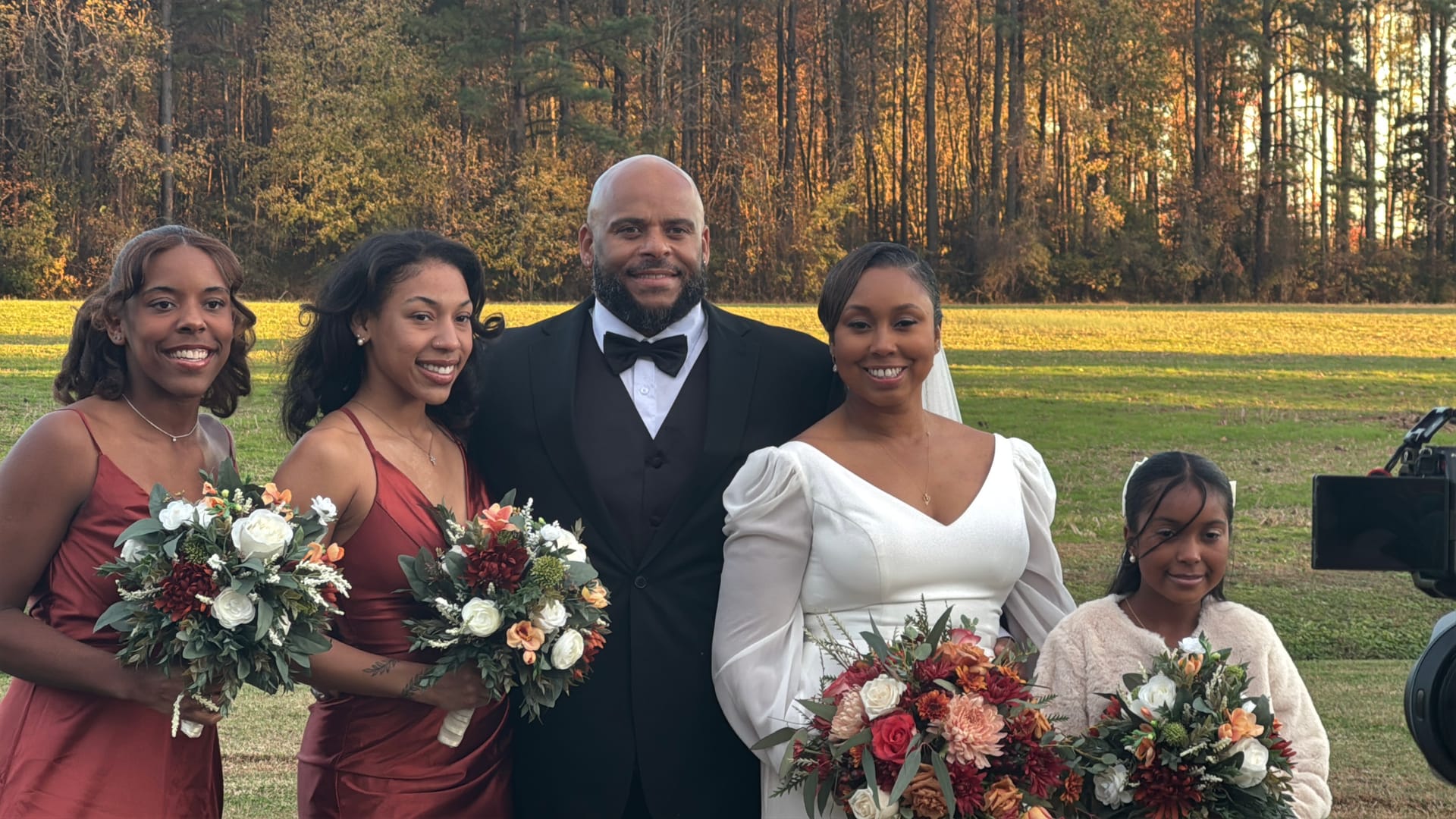 Bride in a white long-sleeve gown and groom in a black tuxedo pose with two bridesmaids in burgundy dresses and a young flower girl holding autumn bouquets against a golden-hour farmland backdrop at Scarborough House.