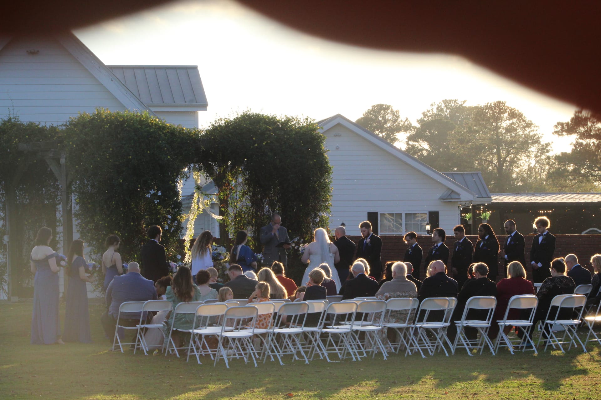 Wedding guests seated on white folding chairs watch a bride and groom exchange vows beneath a lush vine-covered arch on the ceremony lawn at Scarborough House during golden hour.