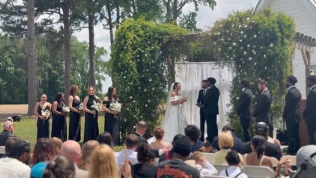 Bride and groom exchange vows beneath a lush greenery and white floral arch on the ceremony lawn at Scarborough House, flanked by bridesmaids in black gowns and groomsmen in suits before a seated outdoor audience.