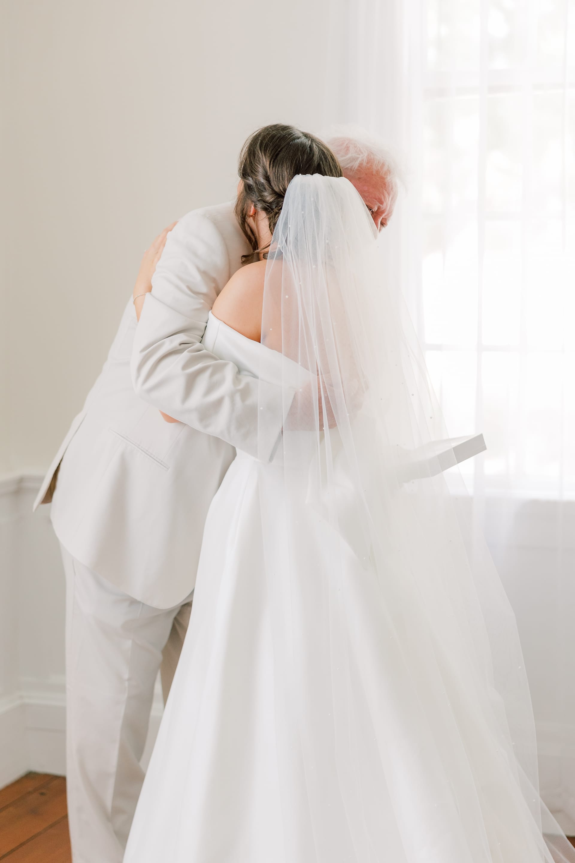 A bride in a white gown and veil shares a heartfelt embrace with a man in a light suit near a bright window inside the Scarborough House bridal suite.