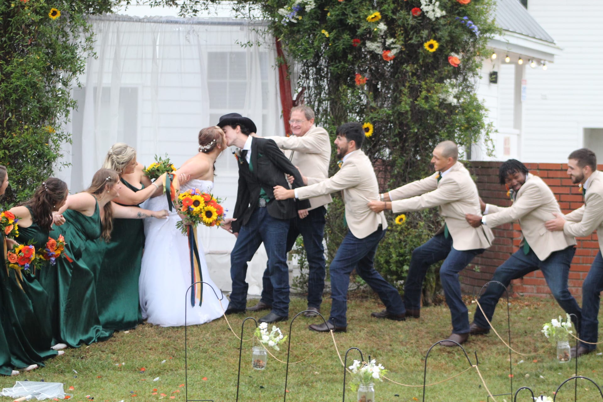 Bride and groom share their first kiss while bridesmaids in emerald green gowns and groomsmen in tan blazers playfully pull them apart on the ceremony lawn at Scarborough House, decorated with sunflowers and string lights.
