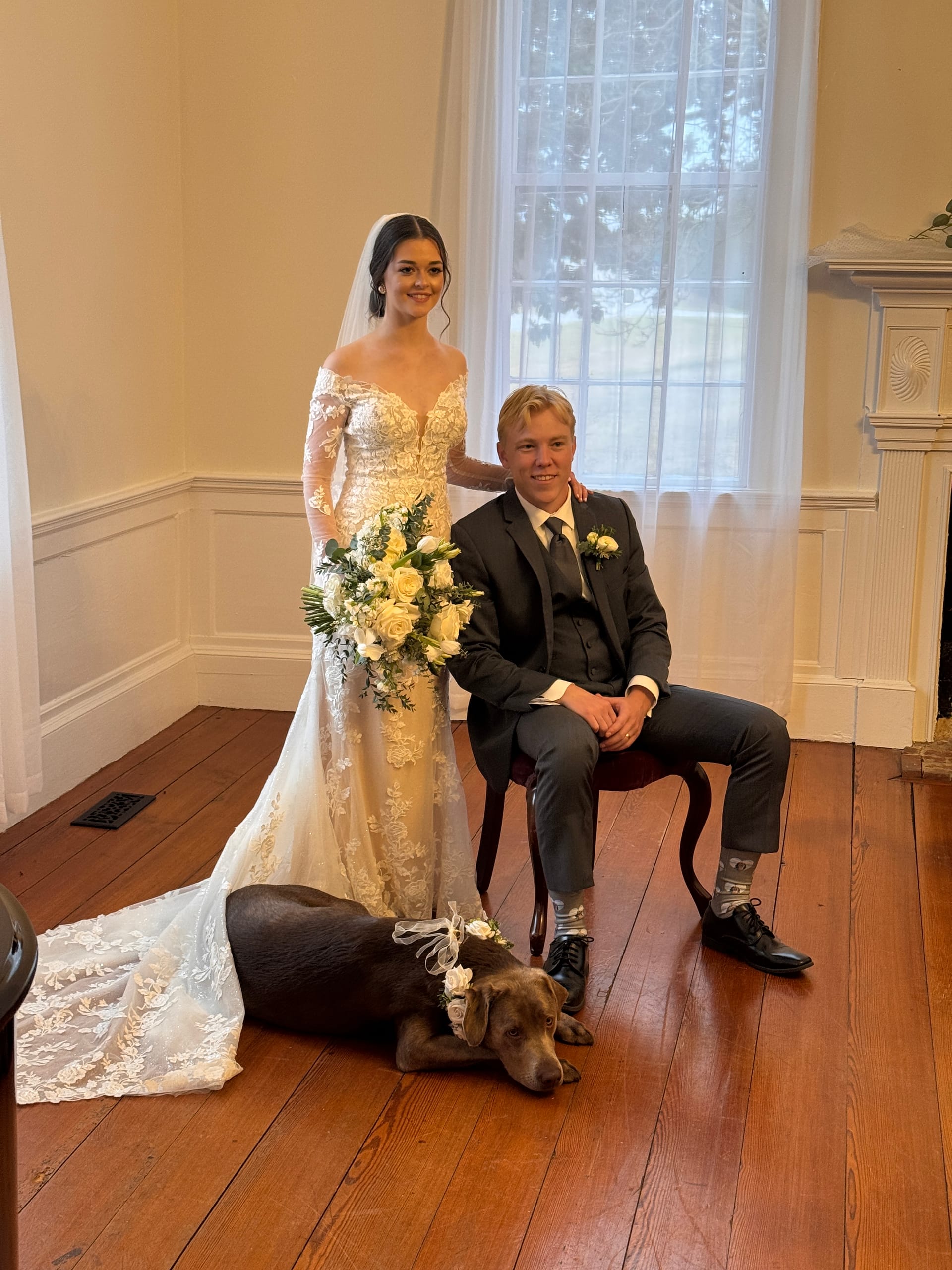 Bride in lace gown holding white rose bouquet stands beside seated groom in dark suit inside a historic manor hall, with their chocolate Labrador wearing a floral crown resting at their feet.