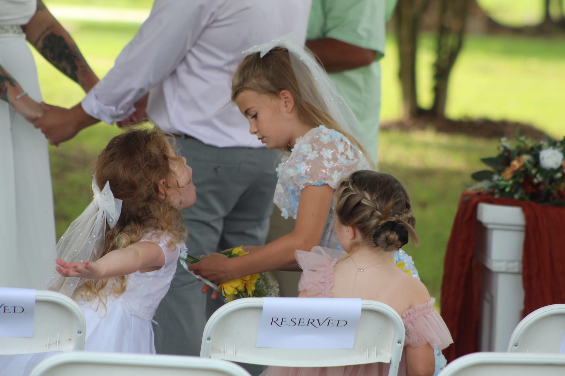 Three young flower girls in white and pastel dresses interact near reserved seating chairs during an outdoor wedding ceremony on the historic estate lawn.