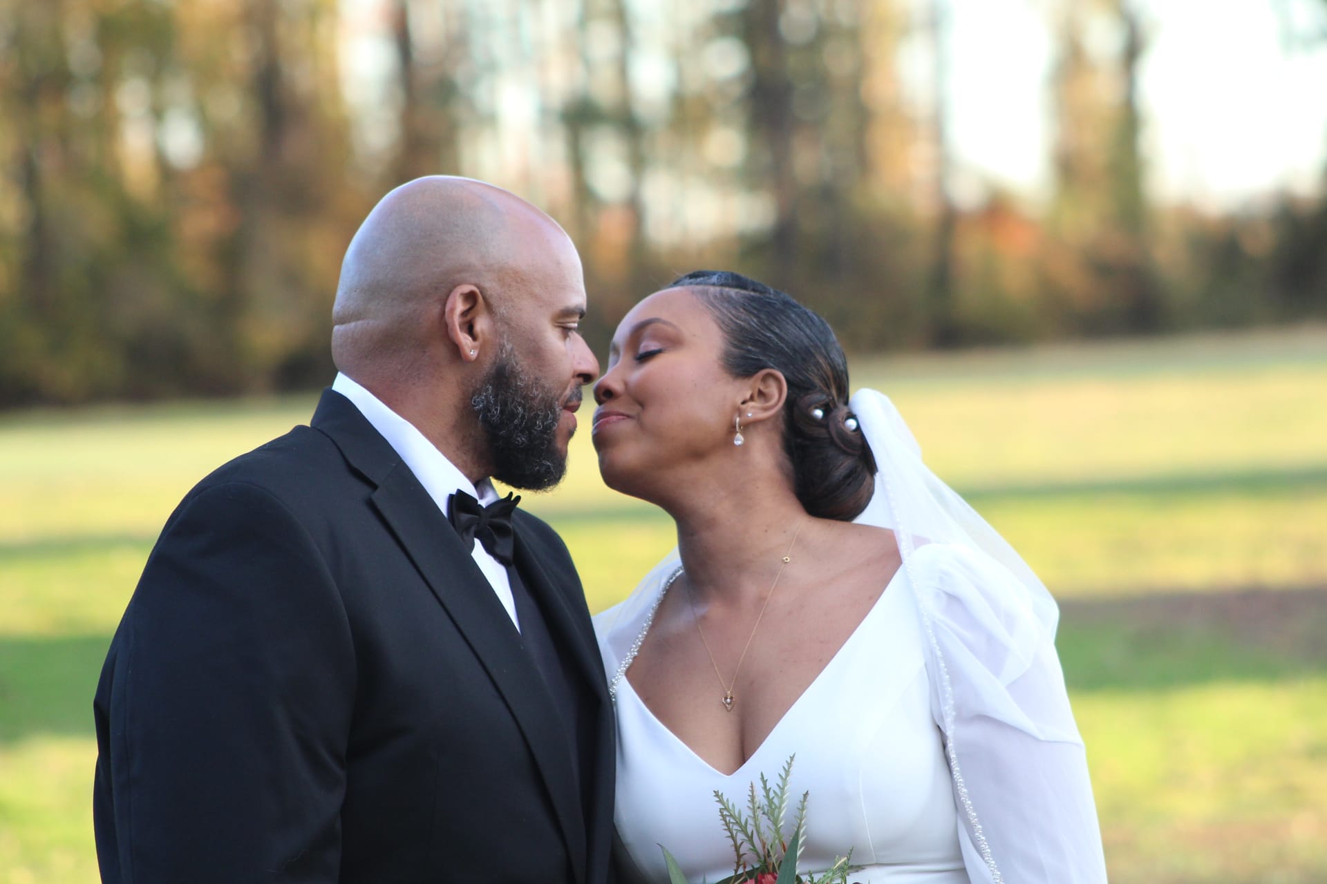 Bride in a white long-sleeve gown and veil leans in for a tender near-kiss with her tuxedo-clad groom against a soft bokeh backdrop of golden autumn trees and open farmland fields at Scarborough House.