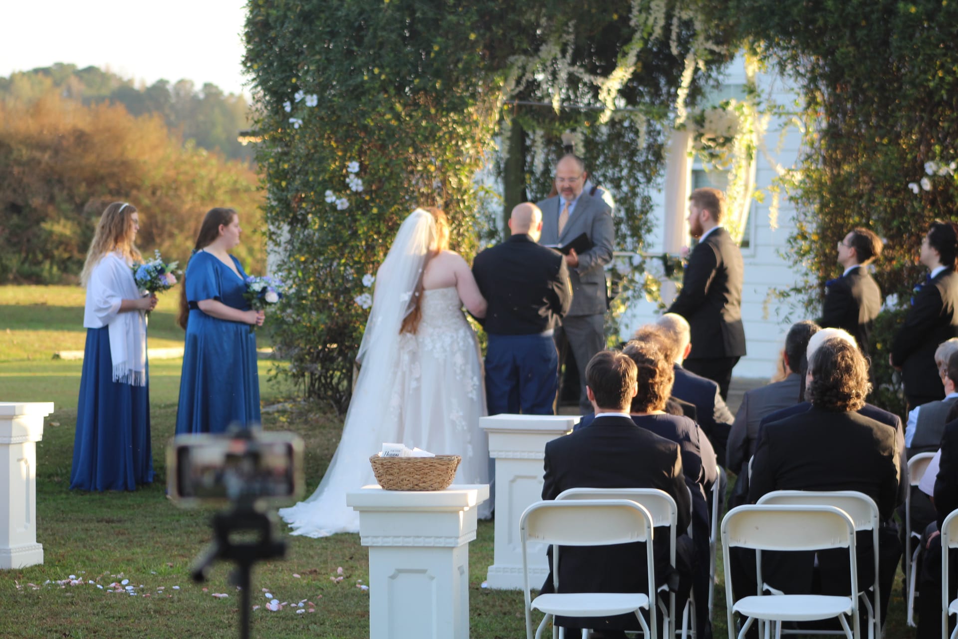 Bride and groom exchange vows before an officiant beneath a lush floral arch on the ceremony lawn at Scarborough House historic estate, with bridesmaids in blue and seated guests in the warm golden afternoon light.