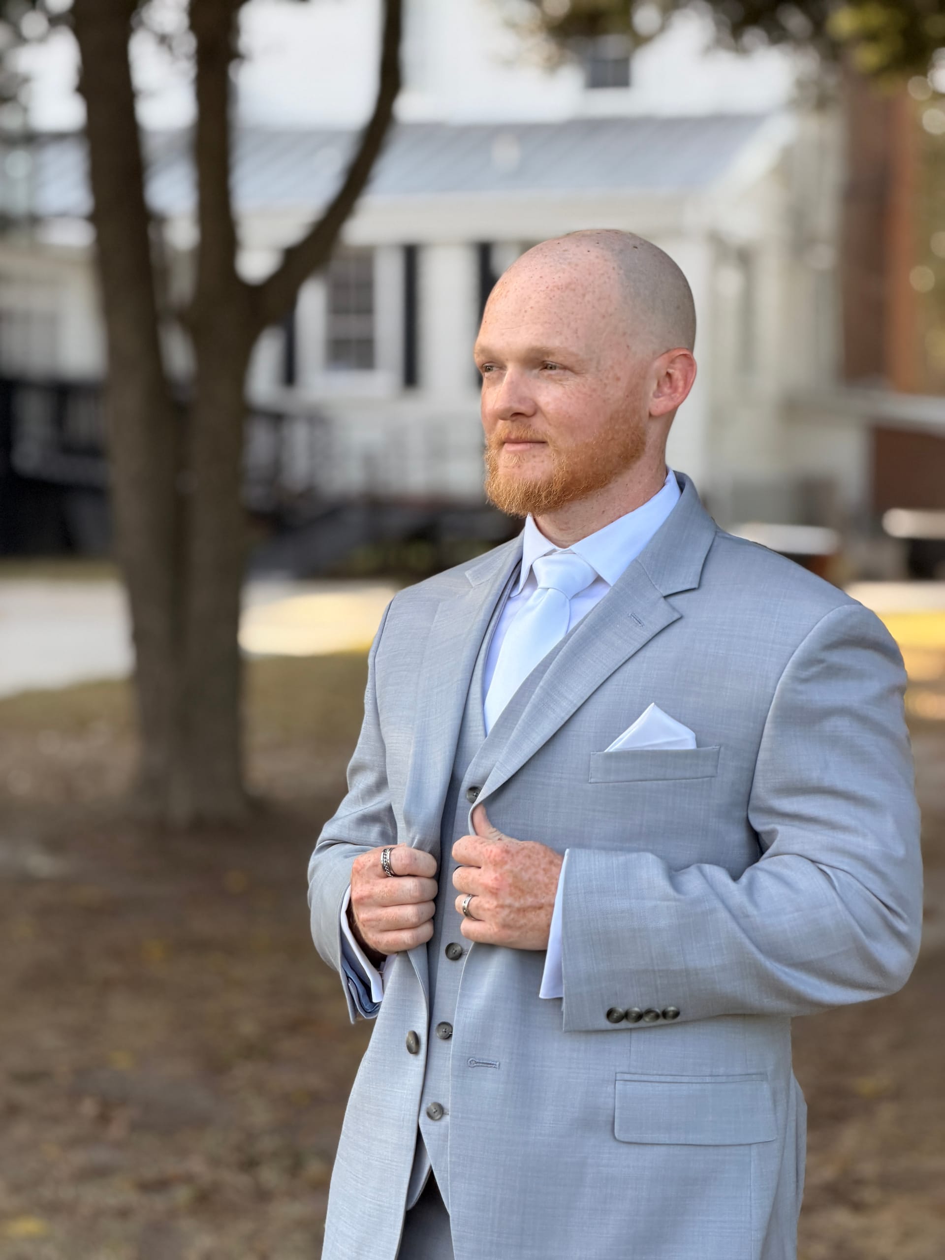 Bald groom with a red beard wearing a light gray three-piece suit and white tie poses confidently outdoors near an oak tree at Scarborough House historic wedding venue.