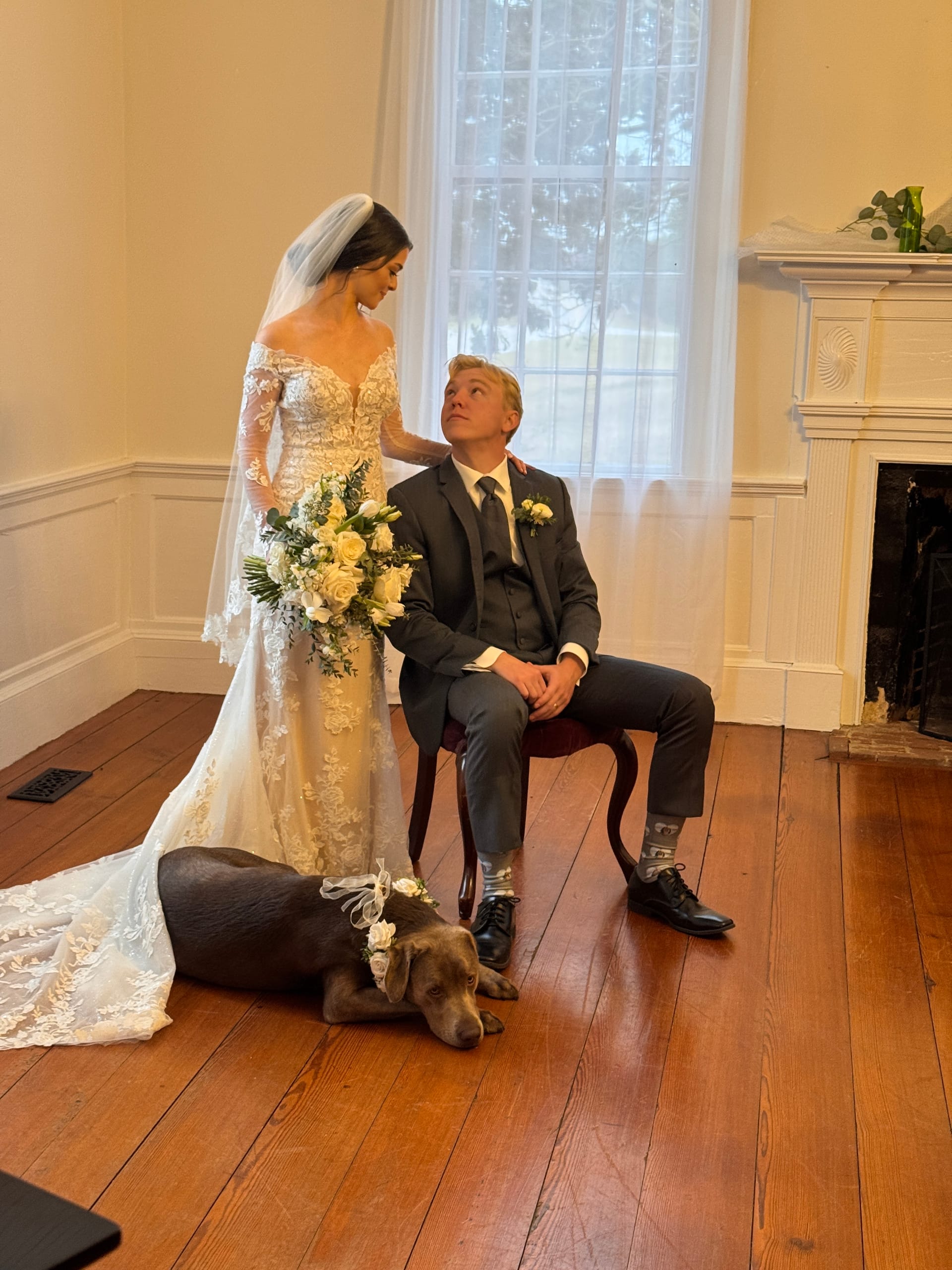 Bride in a lace gown holding a white rose bouquet stands beside her seated groom in a dark suit as their flower-crowned chocolate Labrador rests at their feet inside the historic Scarborough House grand hall.