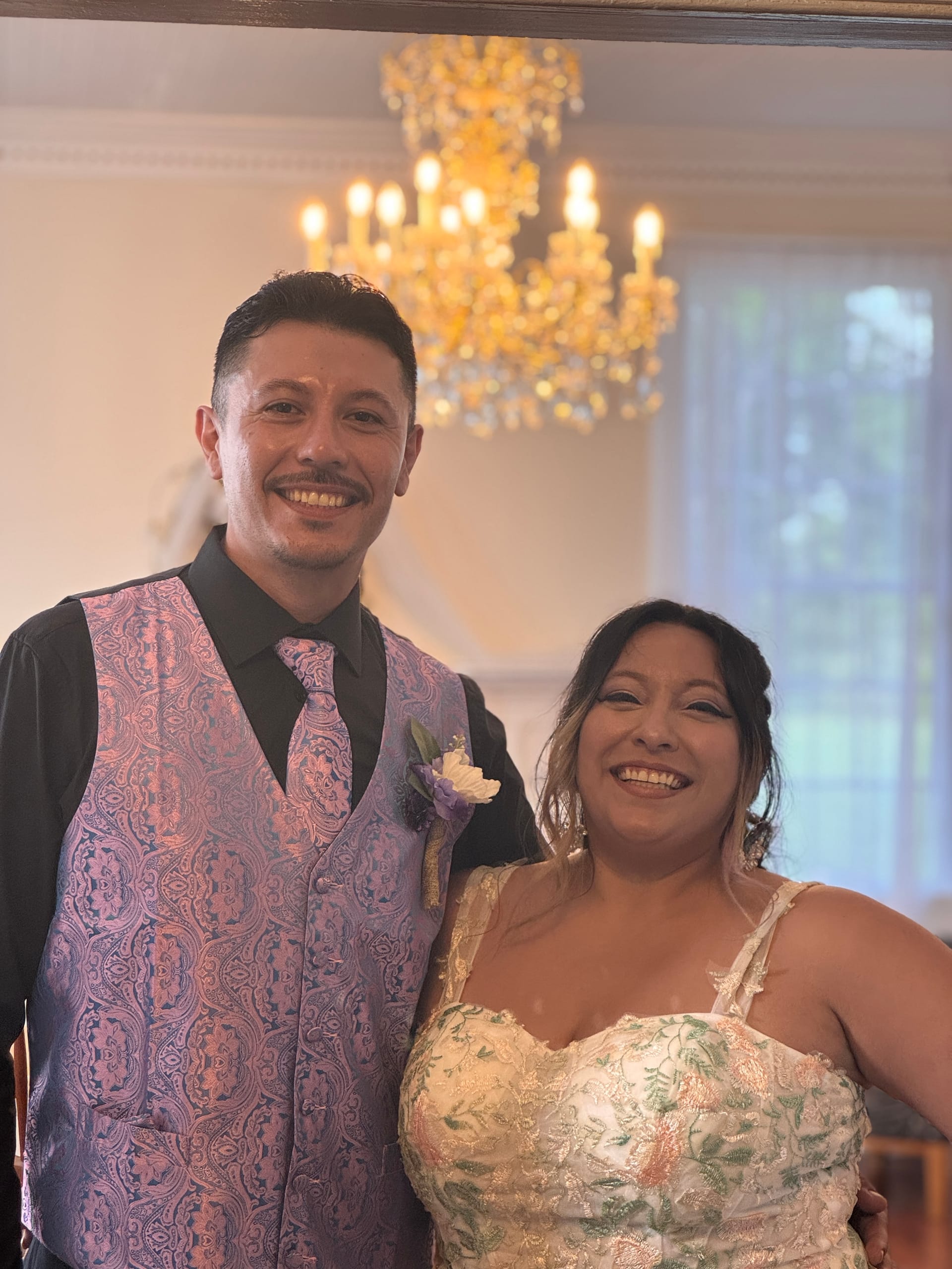 Smiling couple in wedding attire pose together beneath a glowing crystal chandelier inside the historic grand hall at Scarborough House.