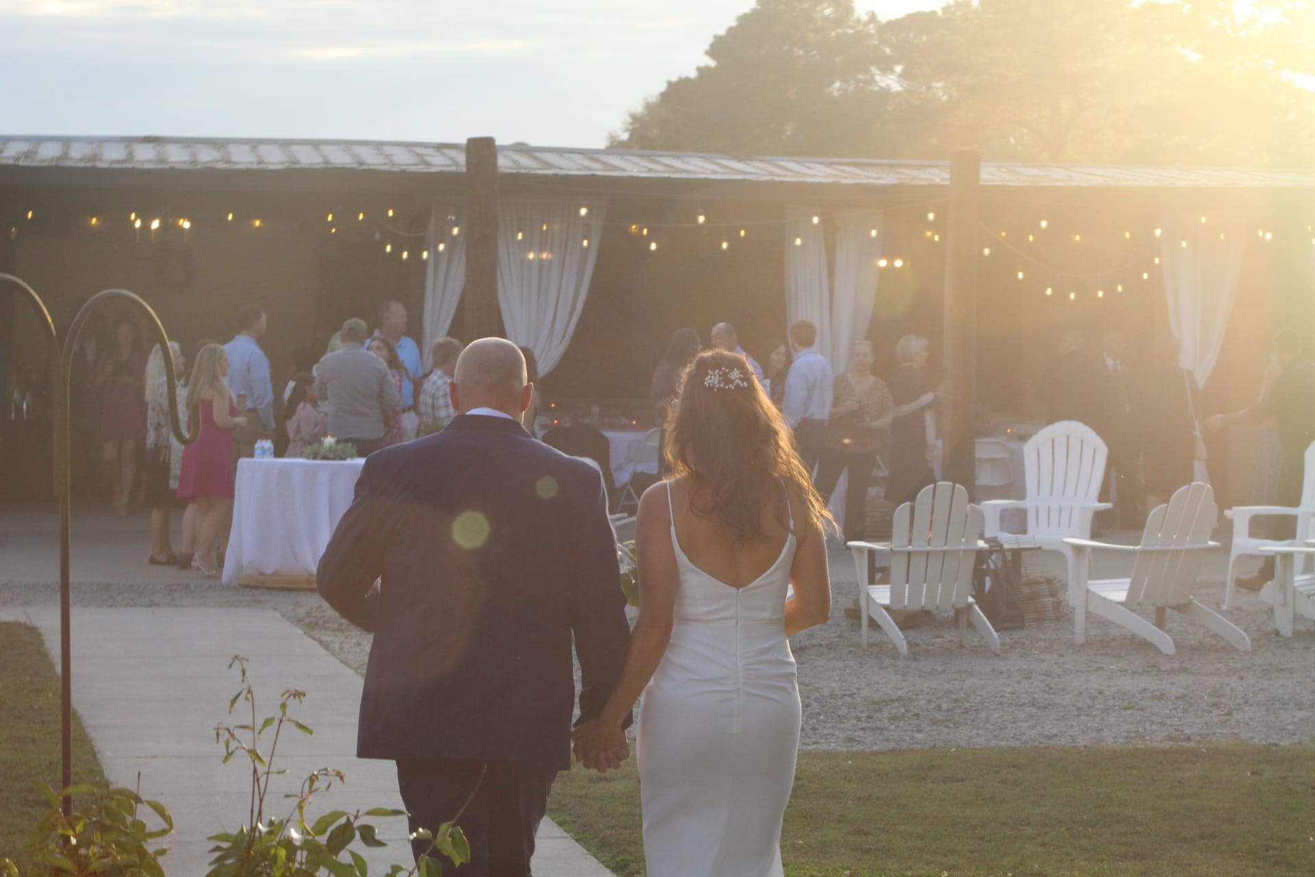 Bride and groom walk hand-in-hand at sunset toward a warmly lit outdoor reception pavilion adorned with string lights and white draping at Scarborough House.