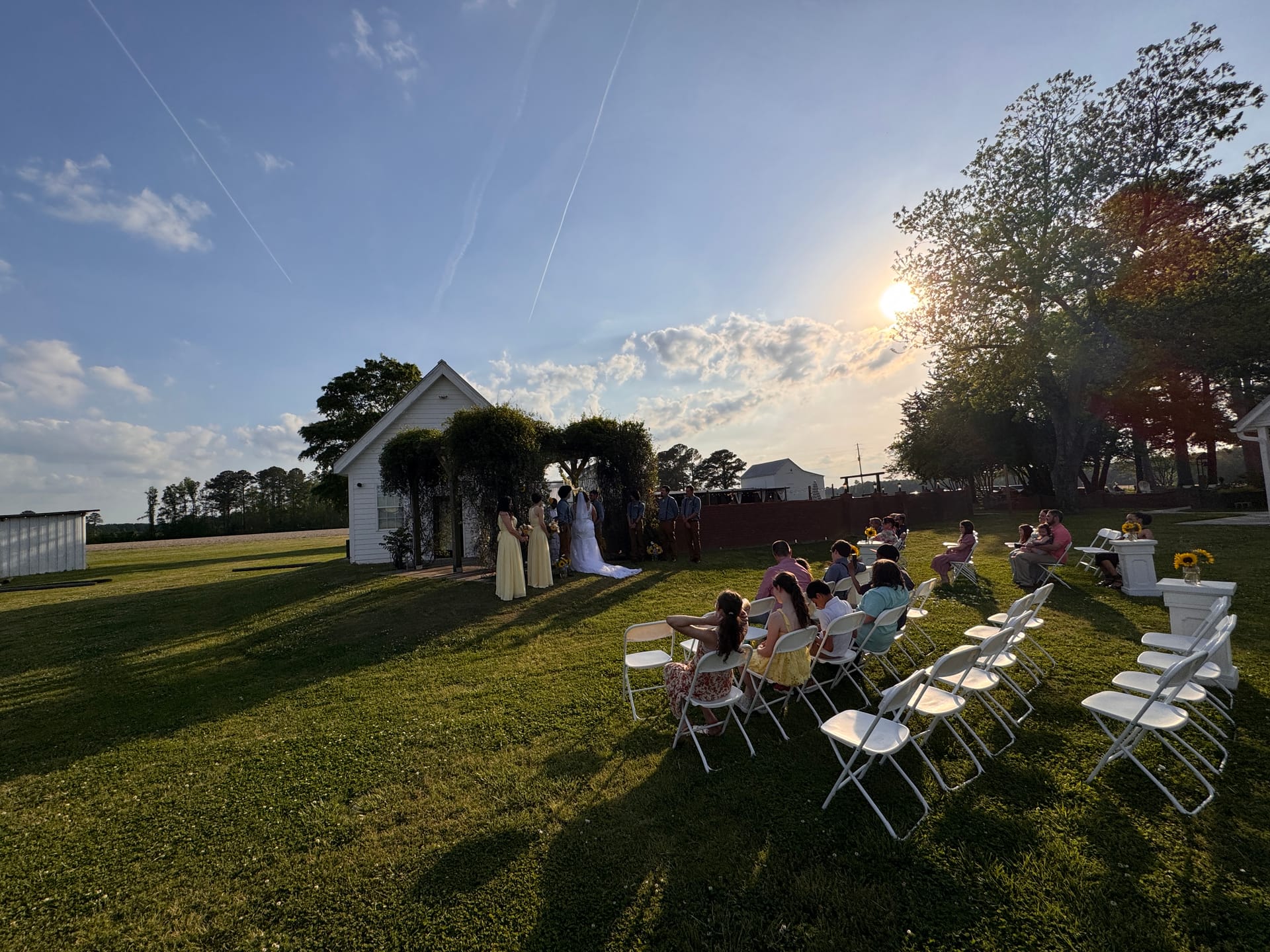 Intimate outdoor wedding ceremony at golden hour on the ceremony lawn at Scarborough House, with guests seated in white folding chairs facing the couple at a vine-covered white chapel arch.