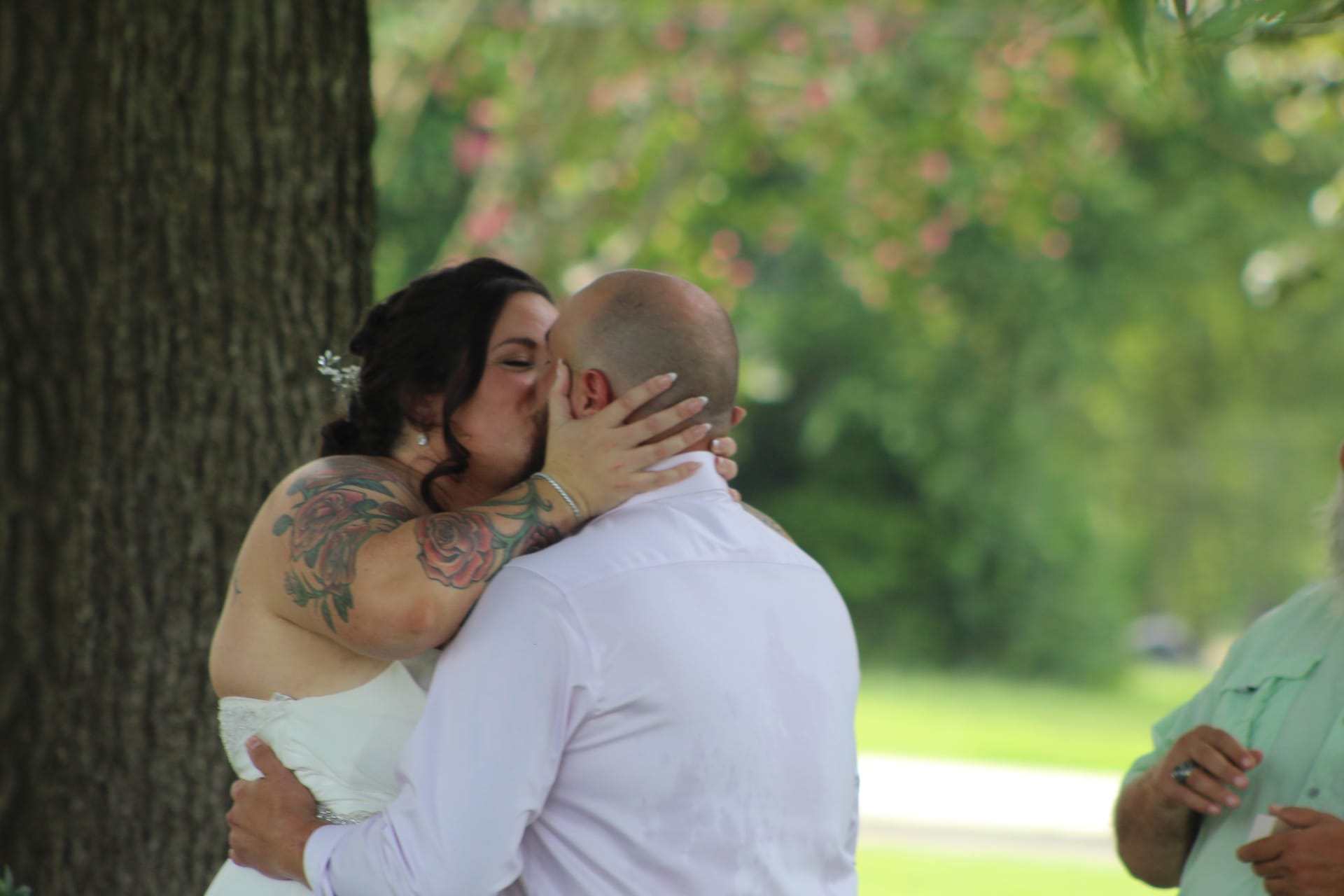 Bride and groom share their first kiss during an outdoor wedding ceremony beneath oak trees at a historic estate venue, with the bride in a white strapless gown and floral tattoo visible on her arm.