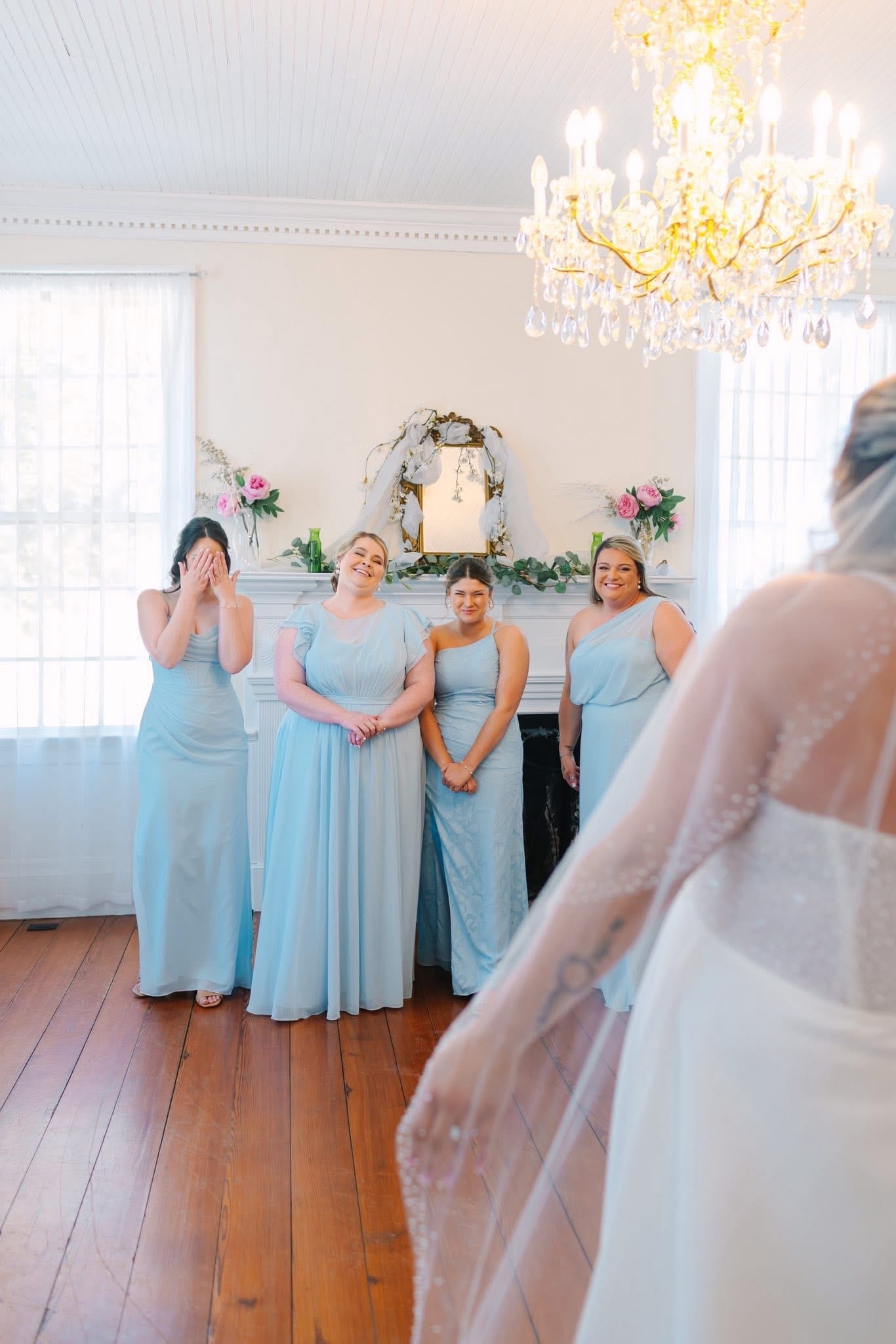 Four bridesmaids in light blue gowns react with joy and tears as the bride in a white gown and veil turns to face them for the first time in a warmly lit historic manor room beneath a crystal chandelier.