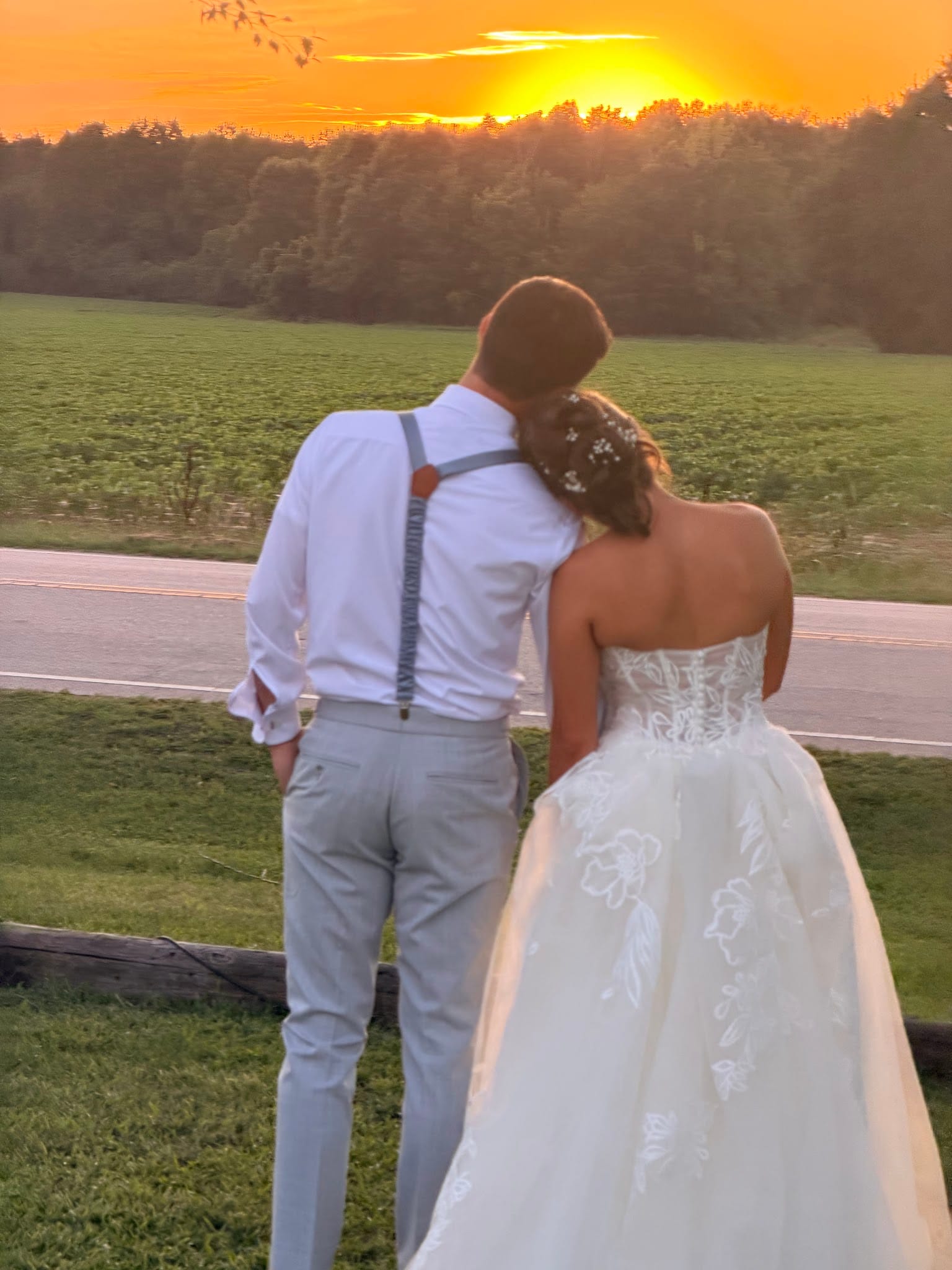 Bride and groom stand together watching a vivid orange sunset over open farmland fields at Scarborough House, the bride resting her head on the grooms shoulder in a tender moment.