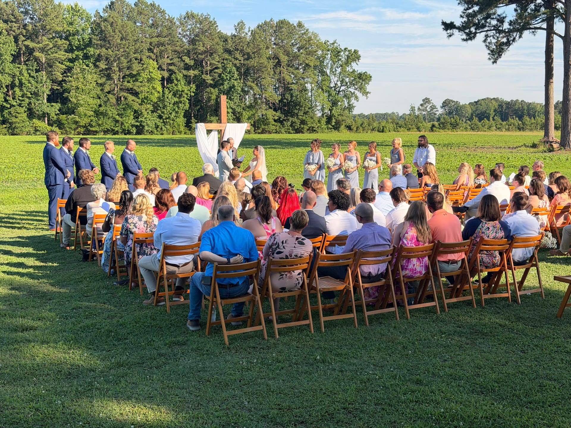 Guests seated in wooden folding chairs watch an outdoor wedding ceremony at a rustic farmland venue with a draped wooden cross altar and lush green fields in the background.
