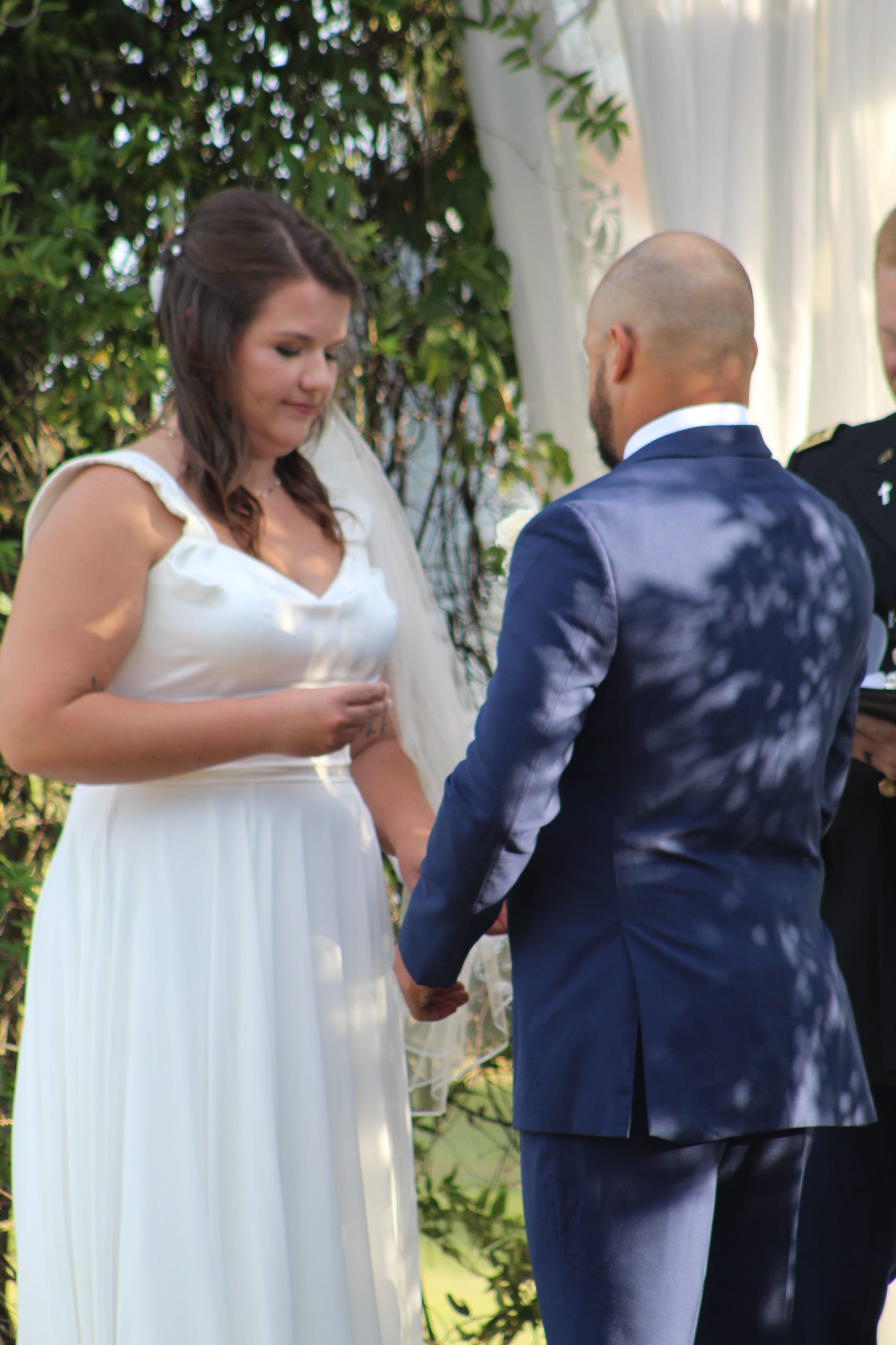 Bride in flowing white gown and veil holds hands with groom in navy suit during an outdoor wedding ceremony surrounded by lush greenery at Scarborough House.