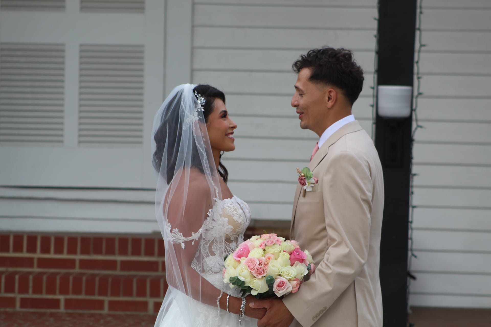 Bride in lace gown and veil holding a pink and white rose bouquet smiles at her groom in a champagne suit with floral boutonniere outside the historic Scarborough House estate.