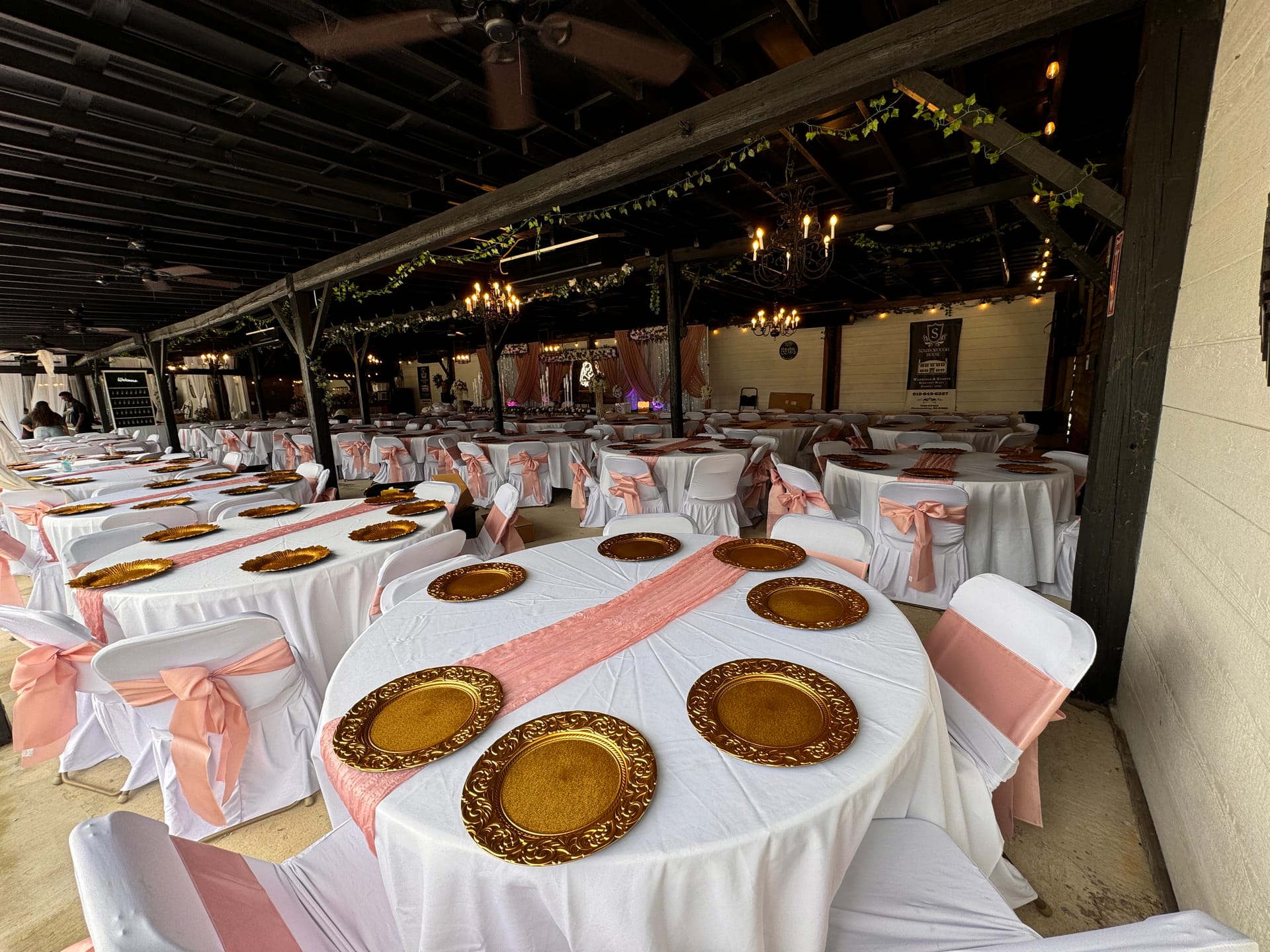 Elegantly decorated reception hall at Scarborough House featuring round tables with white linens, gold charger plates, blush pink table runners and chair sashes, and warm chandelier lighting beneath a rustic wood-beamed pavilion ceiling.