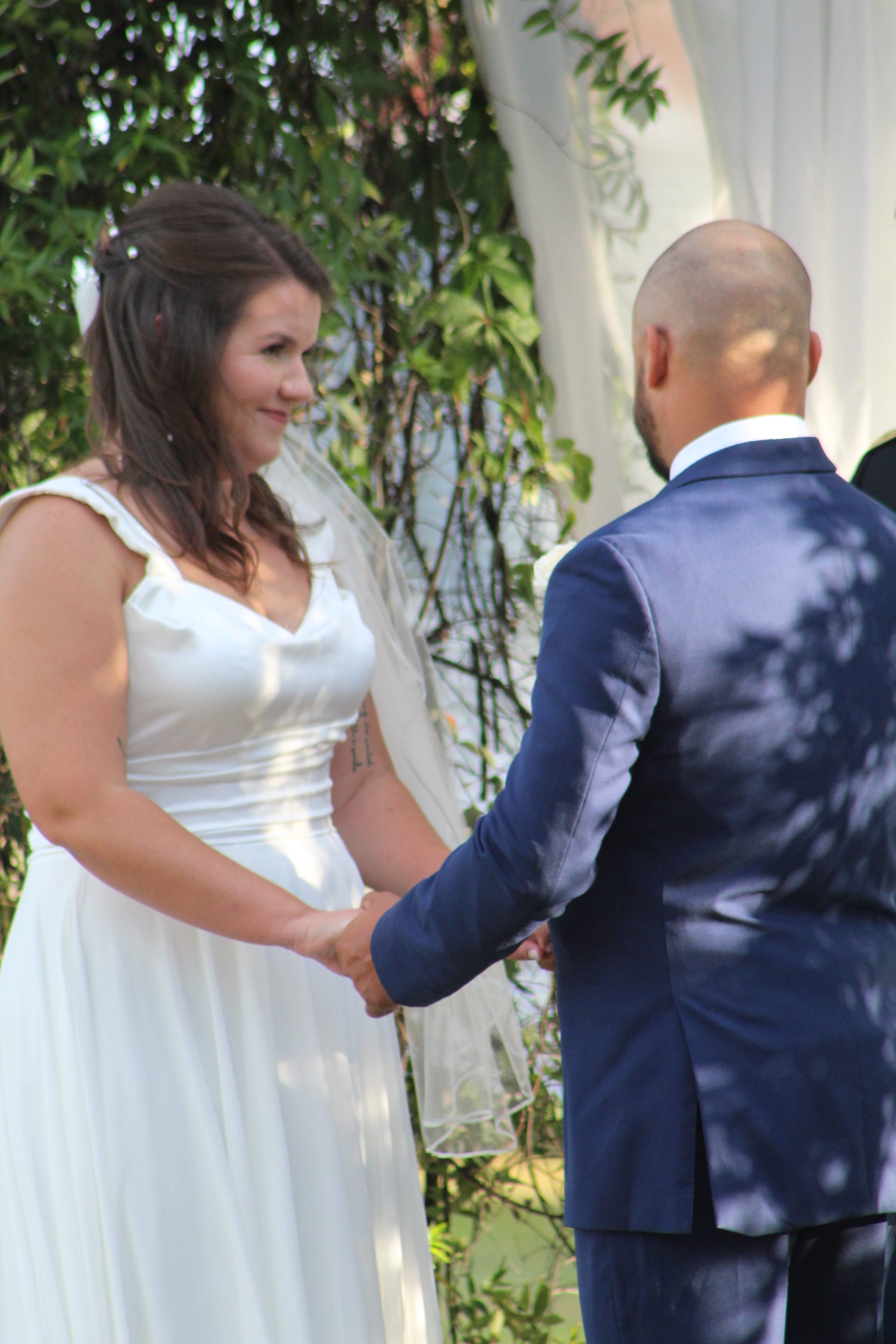 Bride in a flowing white gown smiles warmly at her groom as they hold hands during their outdoor wedding ceremony against a lush green backdrop at Scarborough House.