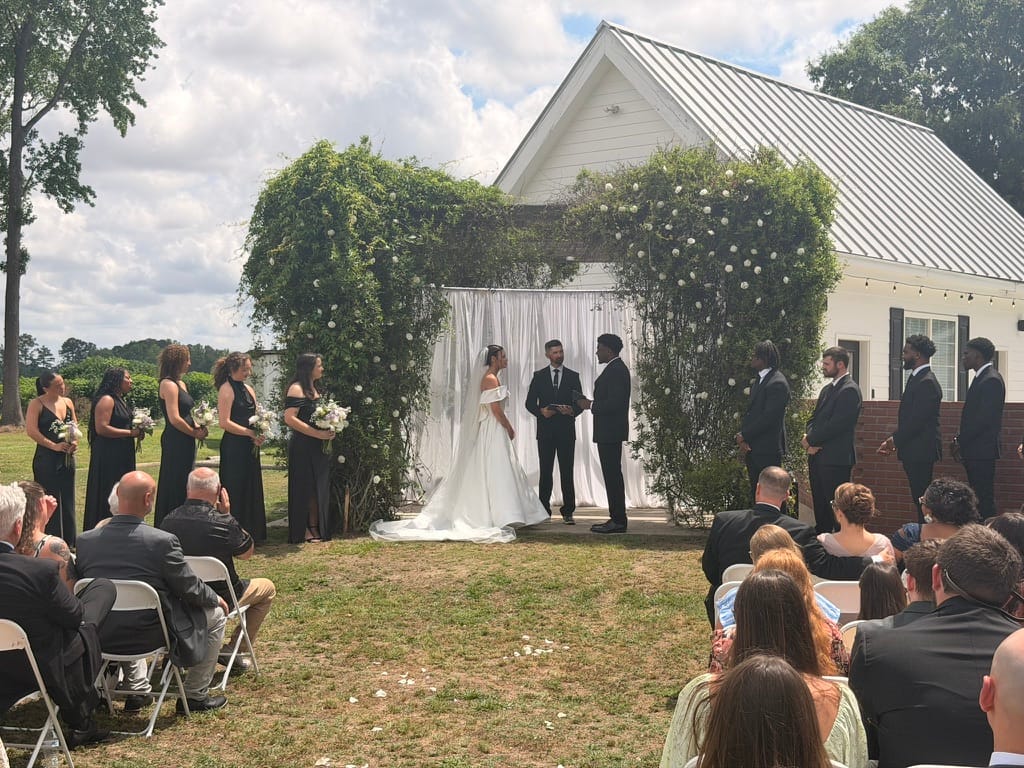 Bride and groom exchange vows beneath an ivy-covered arch adorned with white flowers at an outdoor wedding ceremony at Scarborough House, flanked by bridesmaids in black dresses and groomsmen in suits with seated guests looking on.