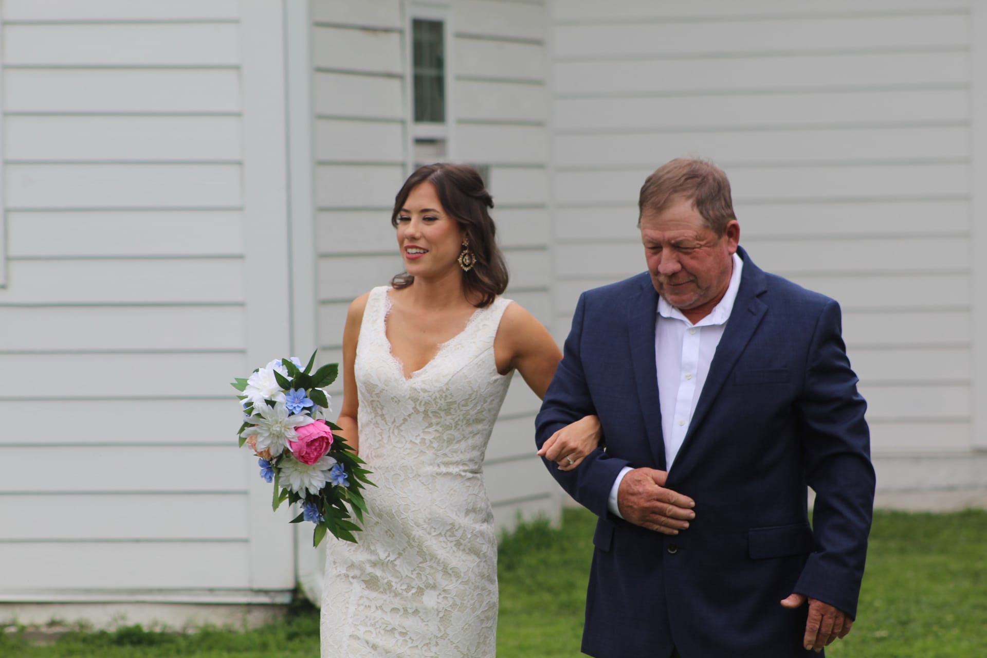 A radiant bride in a lace gown holding a pink and blue bouquet walks arm-in-arm with an older gentleman in a navy suit past the white clapboard exterior of the historic Scarborough House estate.