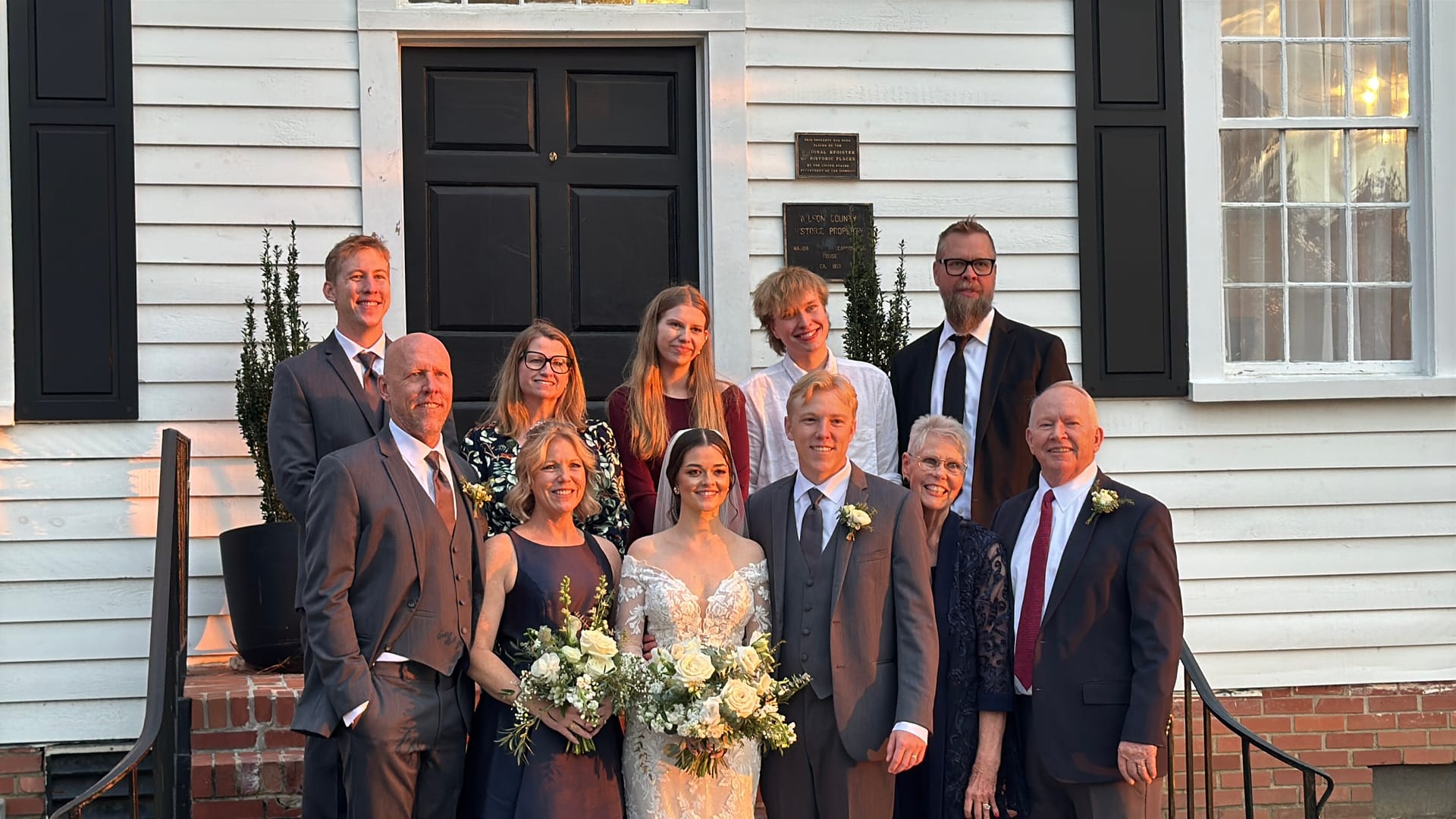 Bride and groom surrounded by smiling family members on the front steps of the 1821 historic white manor house at Scarborough House wedding venue.