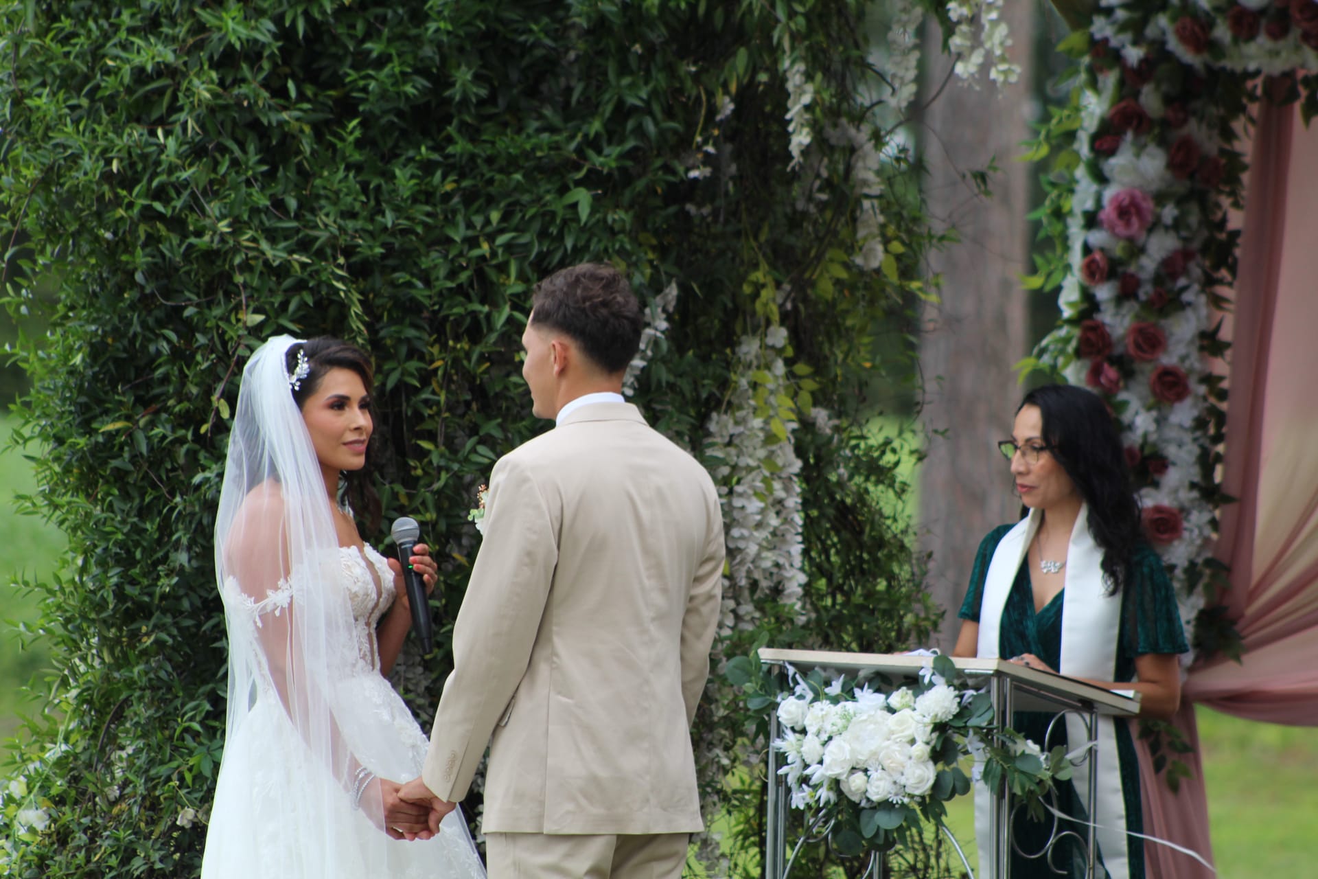 Bride in a lace gown and veil recites her vows into a microphone while holding hands with the groom in a tan suit, officiant standing at a floral-adorned podium beneath a lush greenery arch at Scarborough House.