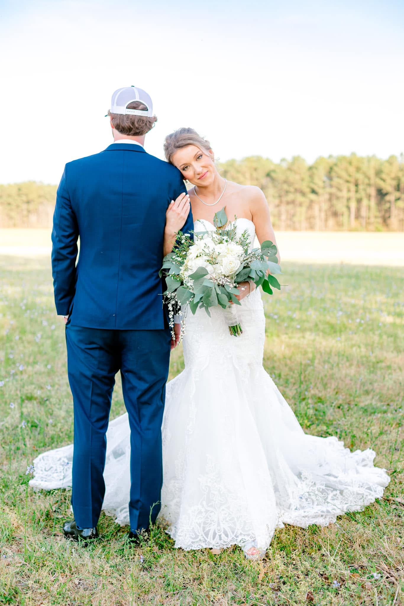Bride in a lace mermaid gown holding a white floral bouquet rests her head on the groom's shoulder as they stand together in an open farmland field during golden hour at Scarborough House.