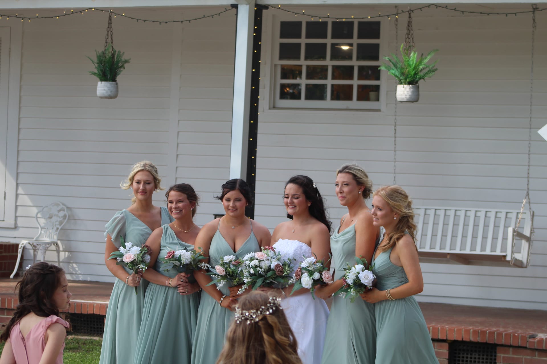 Bride in white gown and five bridesmaids in sage green dresses holding floral bouquets posing together on the wraparound porch of the historic Scarborough House estate.