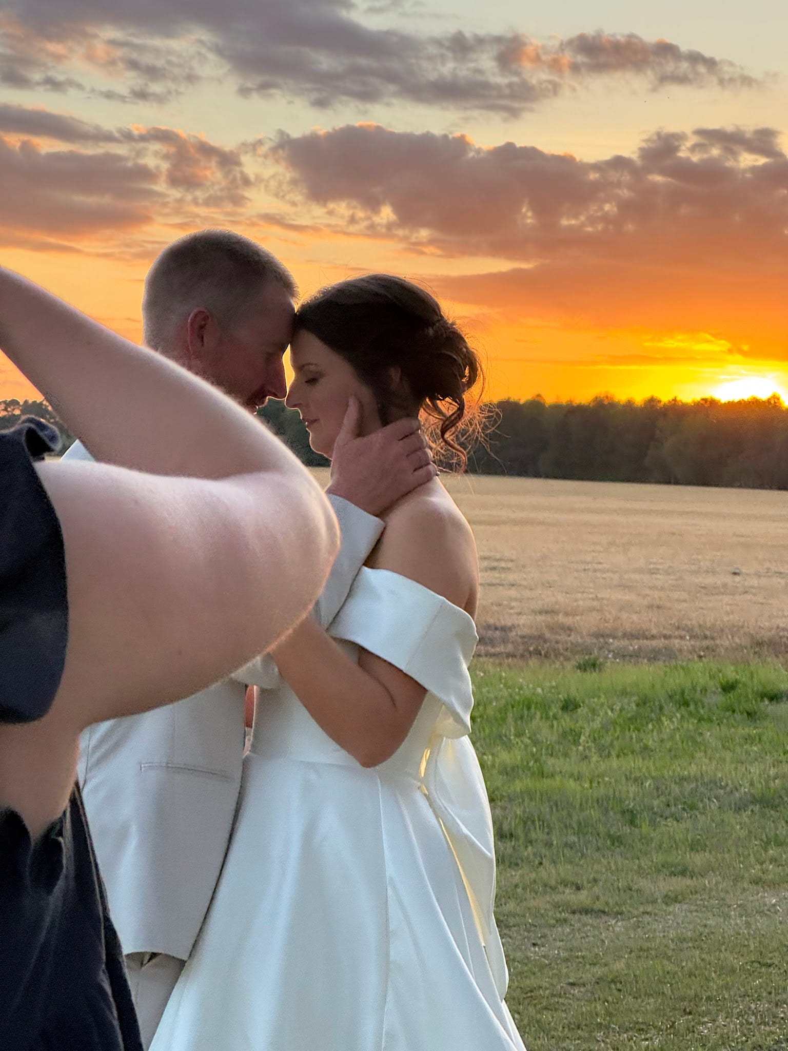 Bride in an off-the-shoulder white gown and groom in a suit share an intimate moment with foreheads nearly touching against a vivid orange and gold sunset over the open farmland fields at Scarborough House.