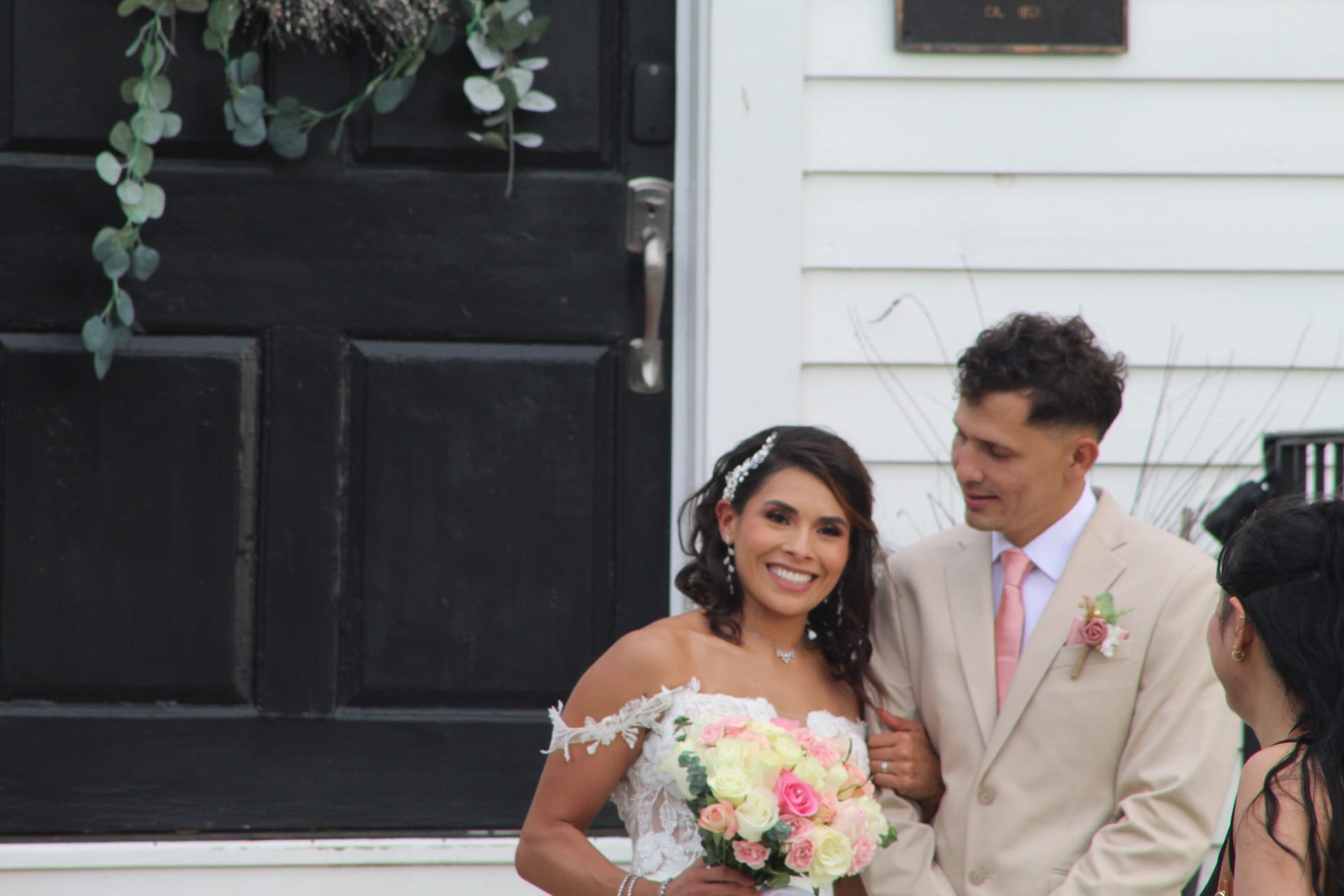 A smiling bride in an off-shoulder lace gown holding a pink and white bouquet stands with her groom in a tan suit beside the historic black door of Scarborough House decorated with a eucalyptus wreath.