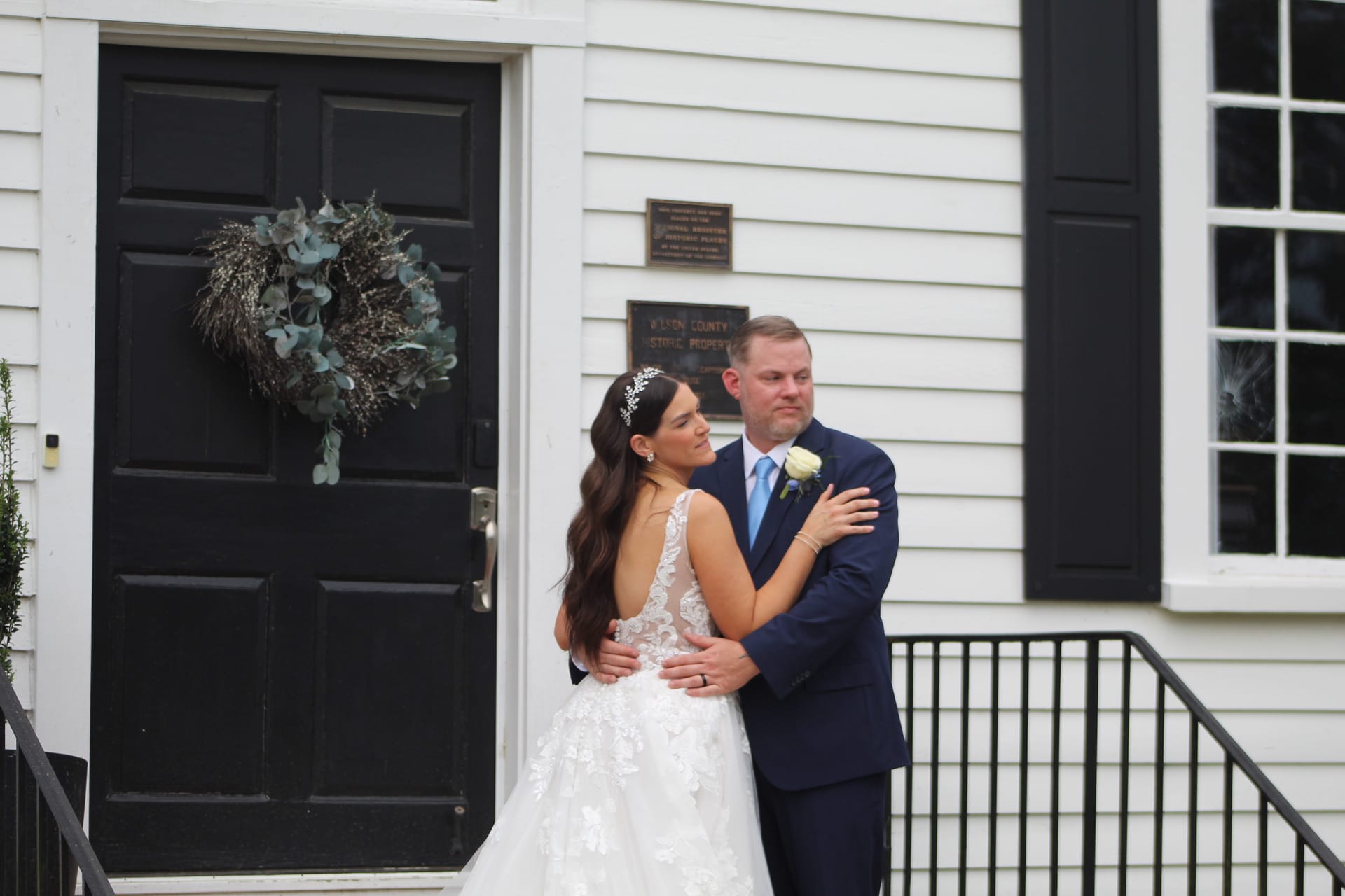 Bride in lace gown and floral headpiece embraces an emotional groom in a navy suit on the front steps of the 1821 historic Scarborough House manor.