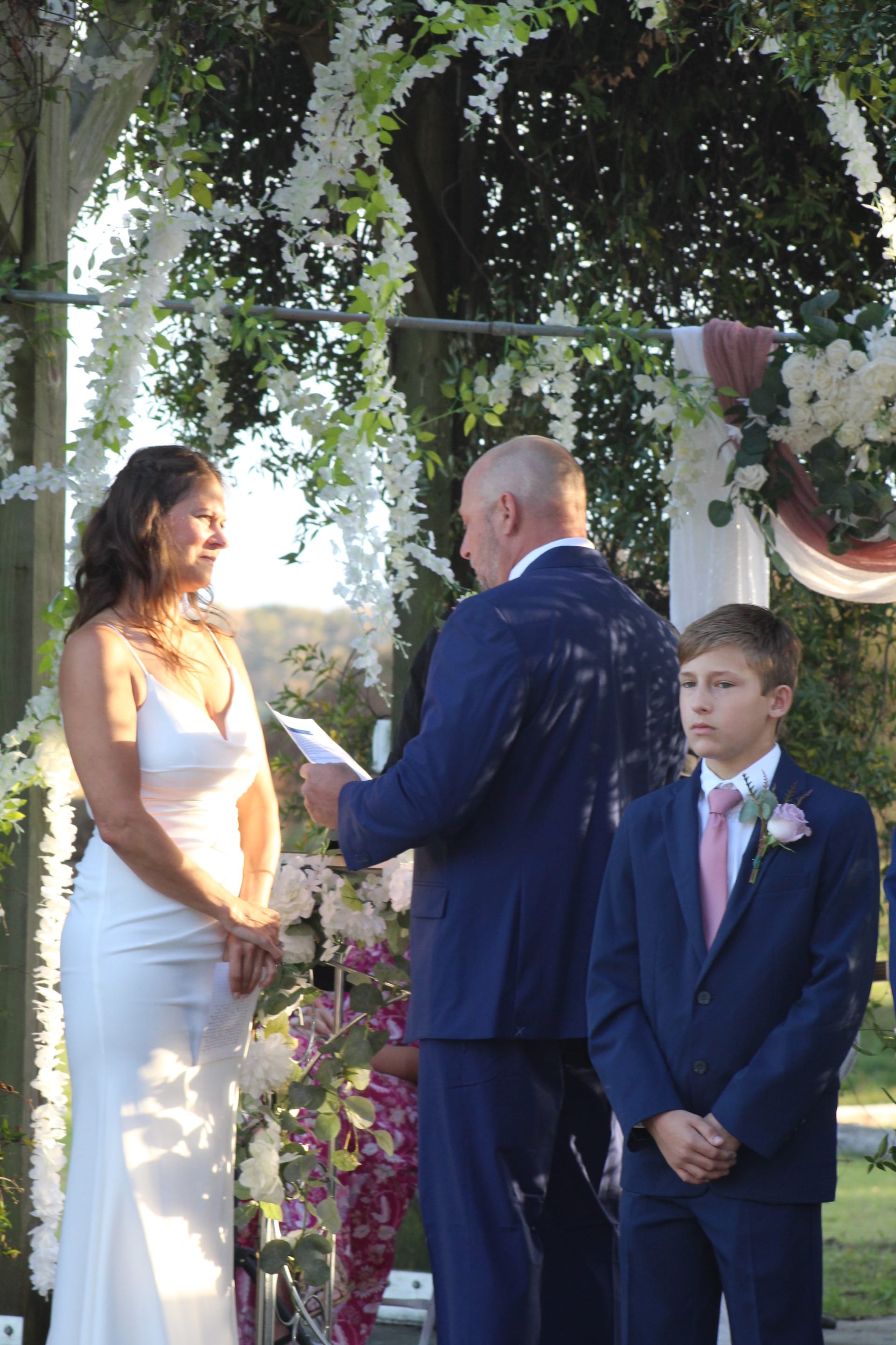 Groom reads vows to a smiling bride in a white gown beneath a lush floral arch on the ceremony lawn at Scarborough House, with a young groomsman standing beside them in a navy suit.