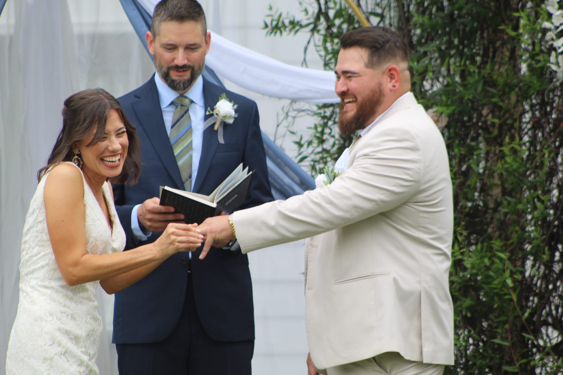 Bride and groom share a joyful laugh while reading vows together as the officiant smiles behind them during an outdoor wedding ceremony at Scarborough House.