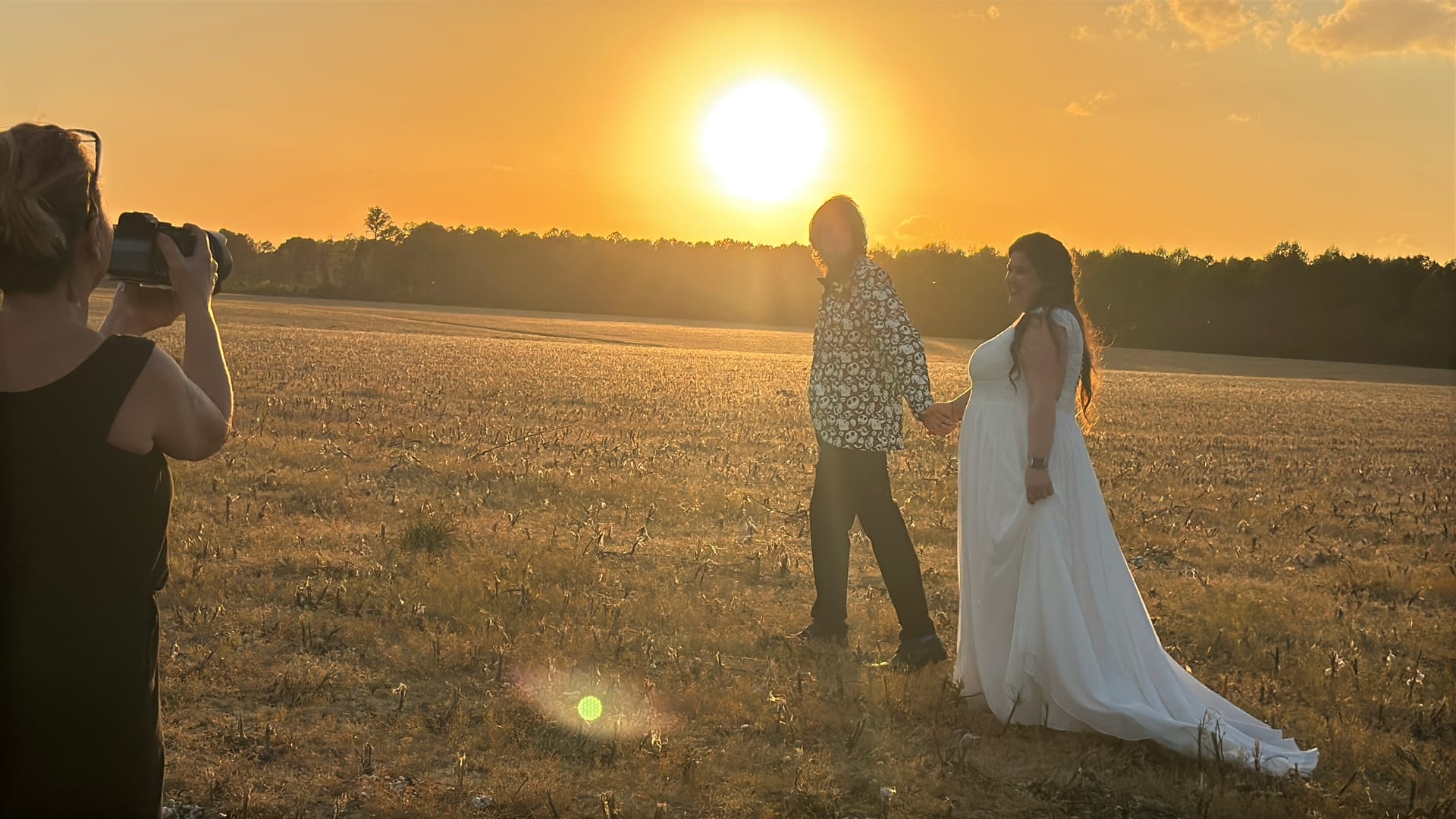 A bride in a flowing white gown and her partner hold hands while walking through a golden farmland field at sunset as a photographer captures the moment at Scarborough House estate.