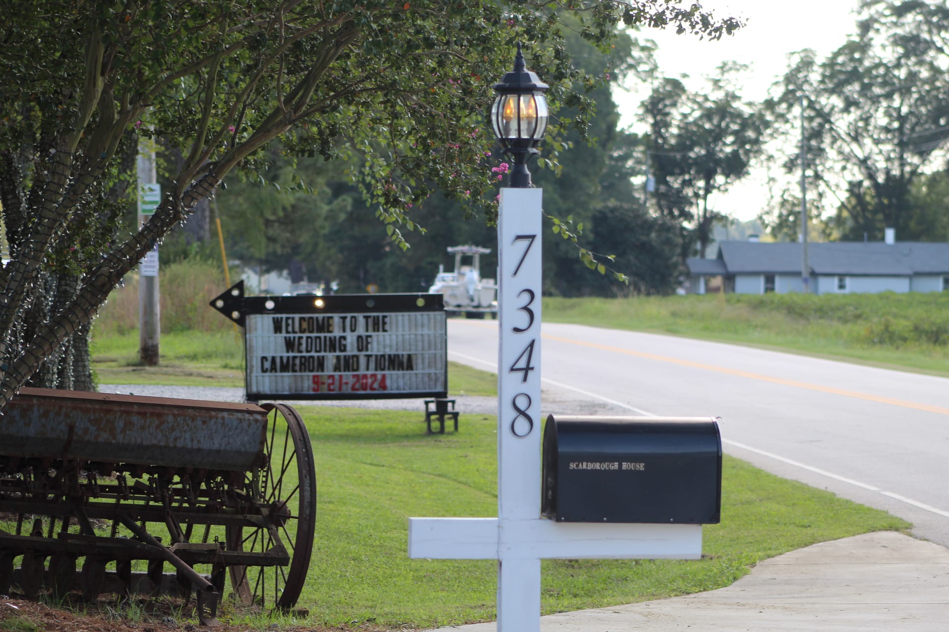 Scarborough House marquee sign welcoming guests to the wedding of Cameron and Tionna on September 21 2024, displayed beside the venue address post at 7348 with a lantern top, vintage farm equipment, and tree-lined road in the background.