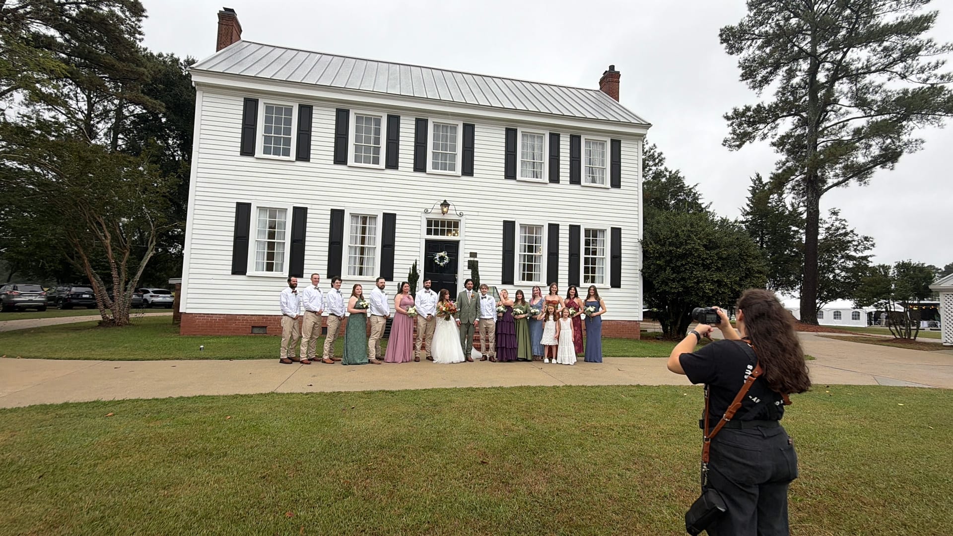 A large wedding party poses in front of the white 1821 Scarborough House manor with black shutters while a photographer captures the group portrait on the ceremony lawn.