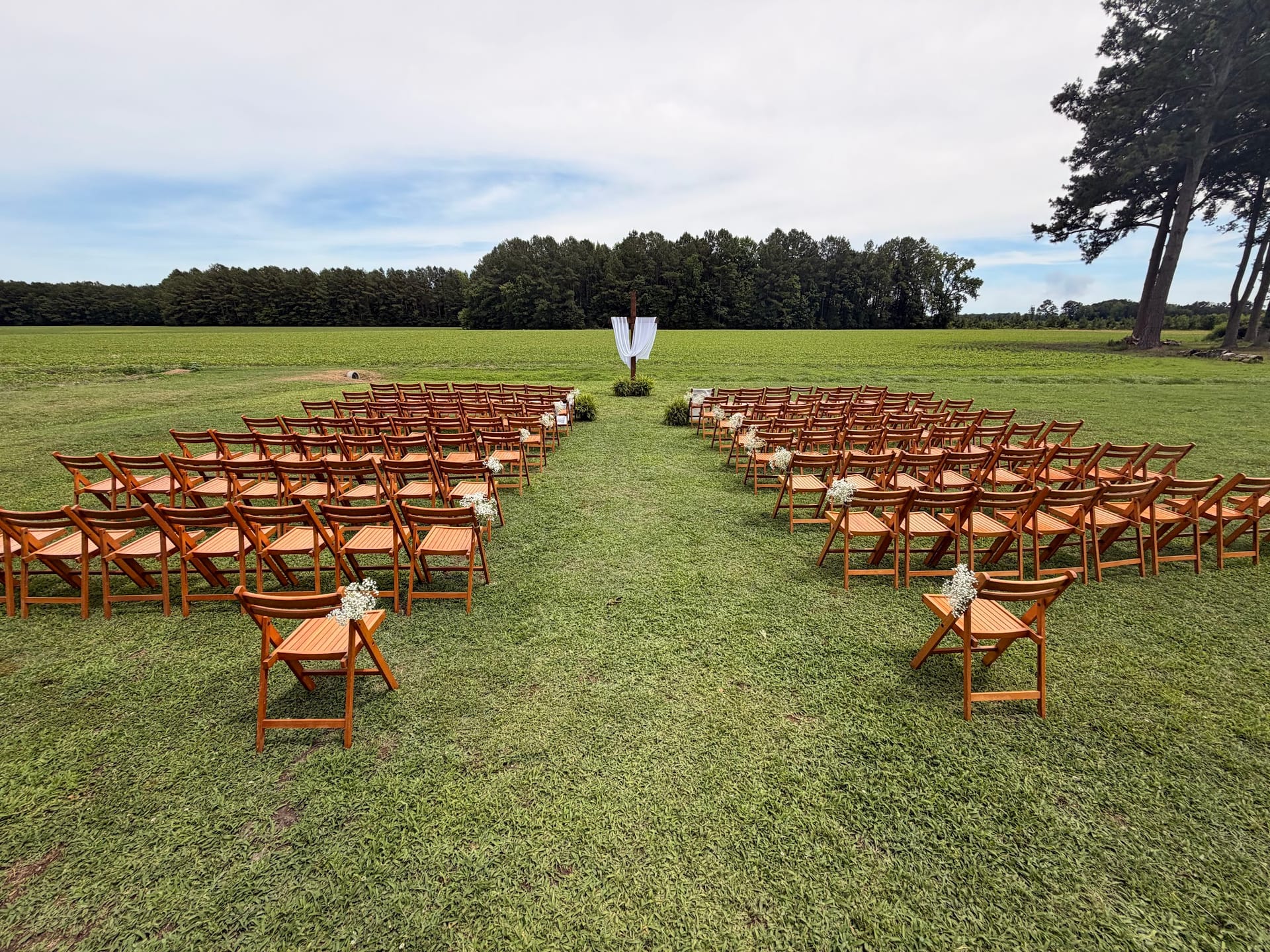 Rows of wooden folding chairs with floral arrangements line a grassy outdoor aisle leading to a draped ceremony arch at Scarborough House wedding venue.
