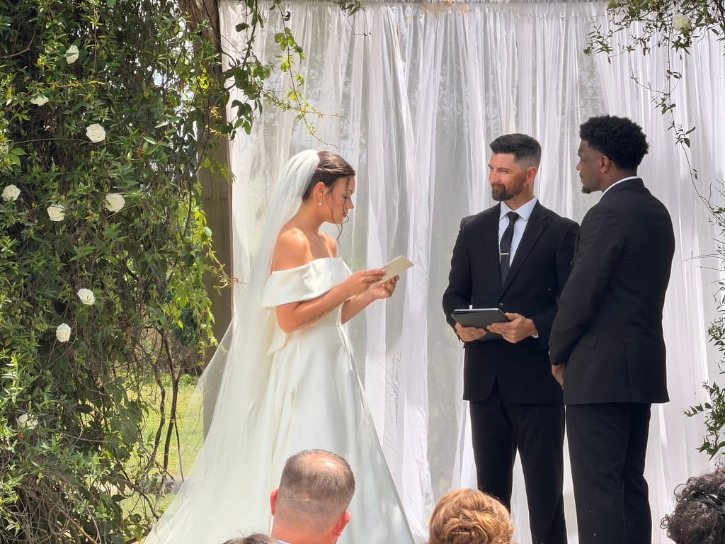 Bride in an off-shoulder white gown reads her vows from a card while the groom in a black suit and officiant look on during an outdoor wedding ceremony at Scarborough House.
