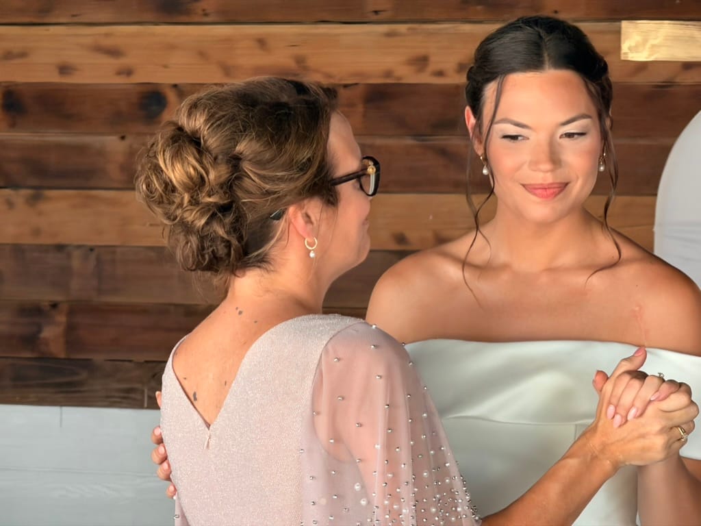 A bride in a white strapless gown smiles warmly while holding hands with a guest in a blush beaded dress inside a rustic wood-paneled room at Scarborough House.