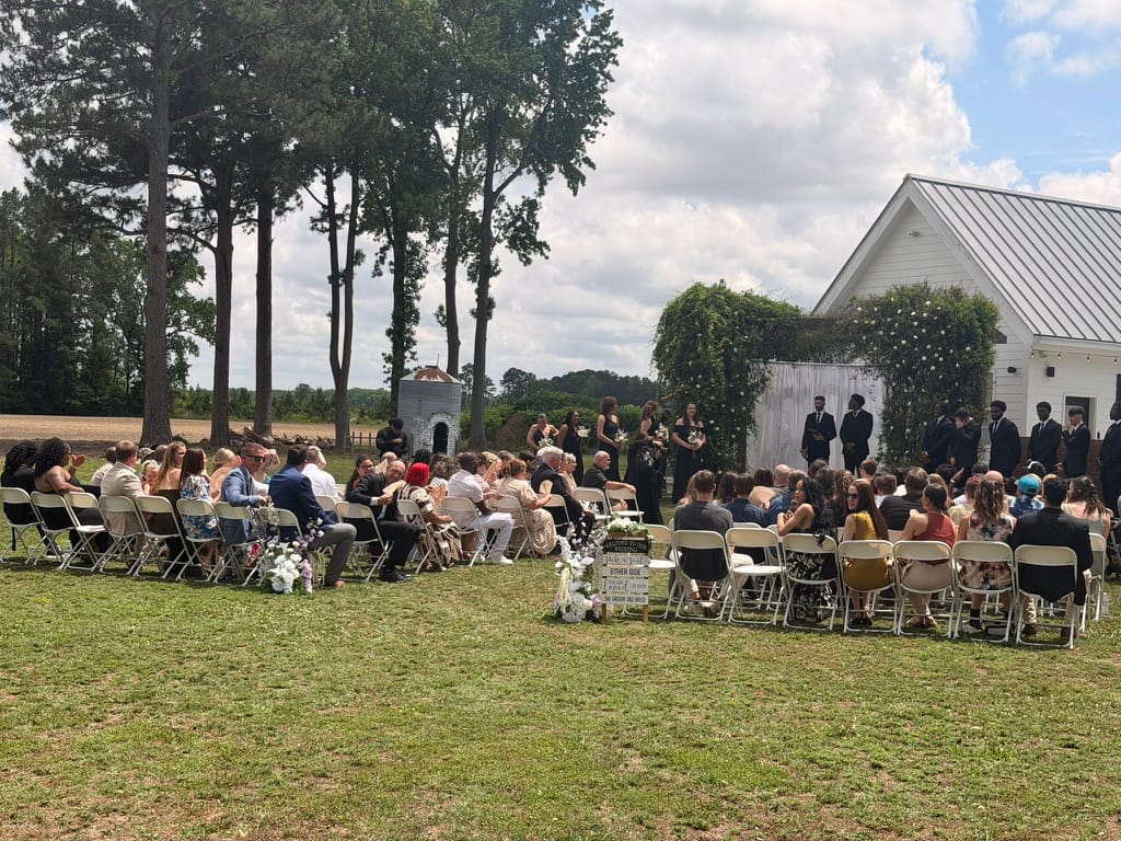 Guests seated on folding chairs during an outdoor wedding ceremony on the sunlit lawn at Scarborough House with a floral-draped white chapel and lush trees in the background.