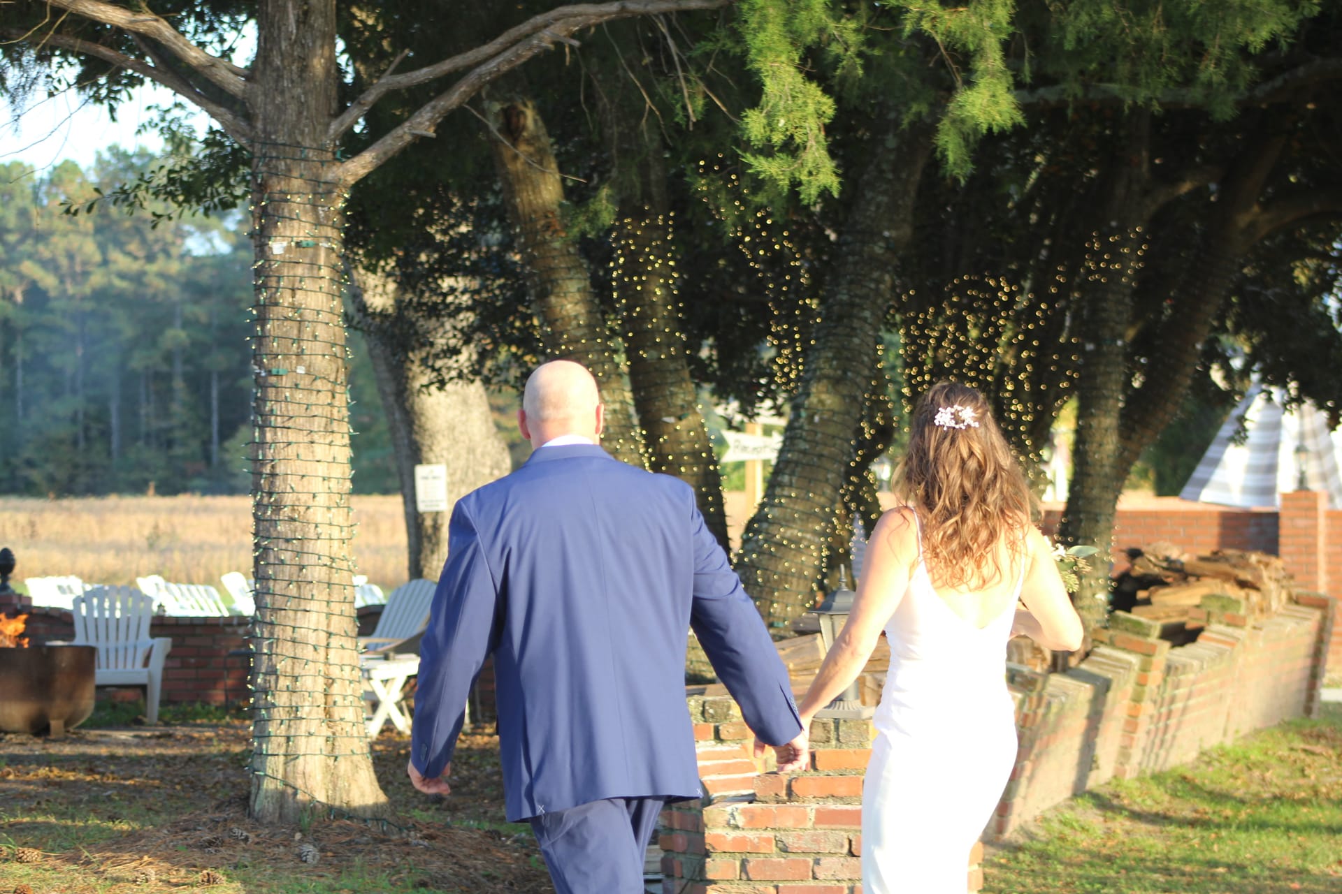 Bride and groom walk hand in hand through a sun-drenched oak tree alley adorned with string lights at Scarborough House estate wedding venue.