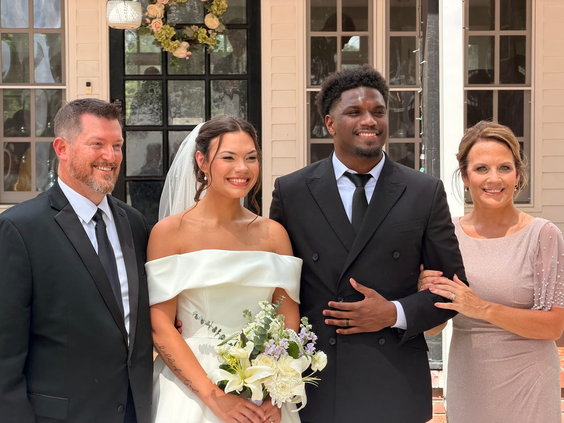 Bride in off-shoulder white gown and groom in black suit pose with two family members in front of the 1821 Scarborough House manor entrance adorned with a floral wreath.